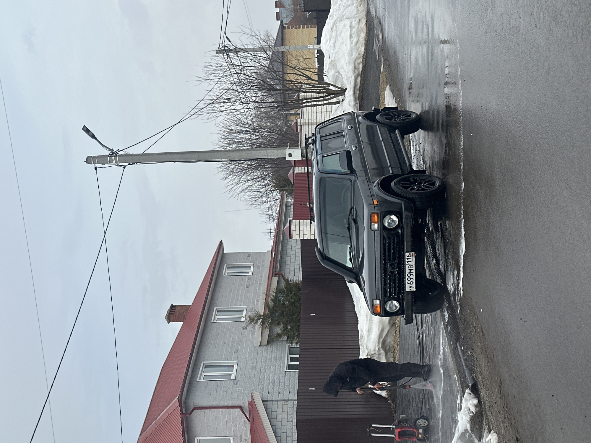 In the city of Kazan, Republic of Tatarstan, Russia, a residential street scene unfolds under an overcast sky during what appears to be late winter or early spring.
A single individual, dressed in a dark hooded jacket and dark pants, is actively operating a red and black portable pressure washer. They are bent over, directing a stream of water from the spray wand onto a wet, dark paved surface adjacent to the street, causing water and foam to splatter.
A dark-colored Lada Niva SUV, equipped with a roof rack and black multi-spoke wheels, is parked on the wet asphalt street, facing slightly towards the left foreground. Its front license plate, "У699МВ 116 RUS," is clearly legible.
To the left, behind a dark brown horizontal slatted fence, stands a two-story house featuring light grey, possibly stone-block or textured stucco, exterior walls and a reddish-orange corrugated metal roof with a brick chimney. White-framed windows are visible on the house. Patches of dirty snow are present on the ground along the fence line and roadside.
In the mid-ground, a tall concrete utility pole with a street light fixture and multiple overhead power lines is prominent. Bare deciduous trees, devoid of leaves, are scattered throughout the background. Other residential structures, including one with a yellow brick exterior, are visible further in the background. The street surface is dark, wet asphalt with visible puddles and areas disturbed by the pressure washing.