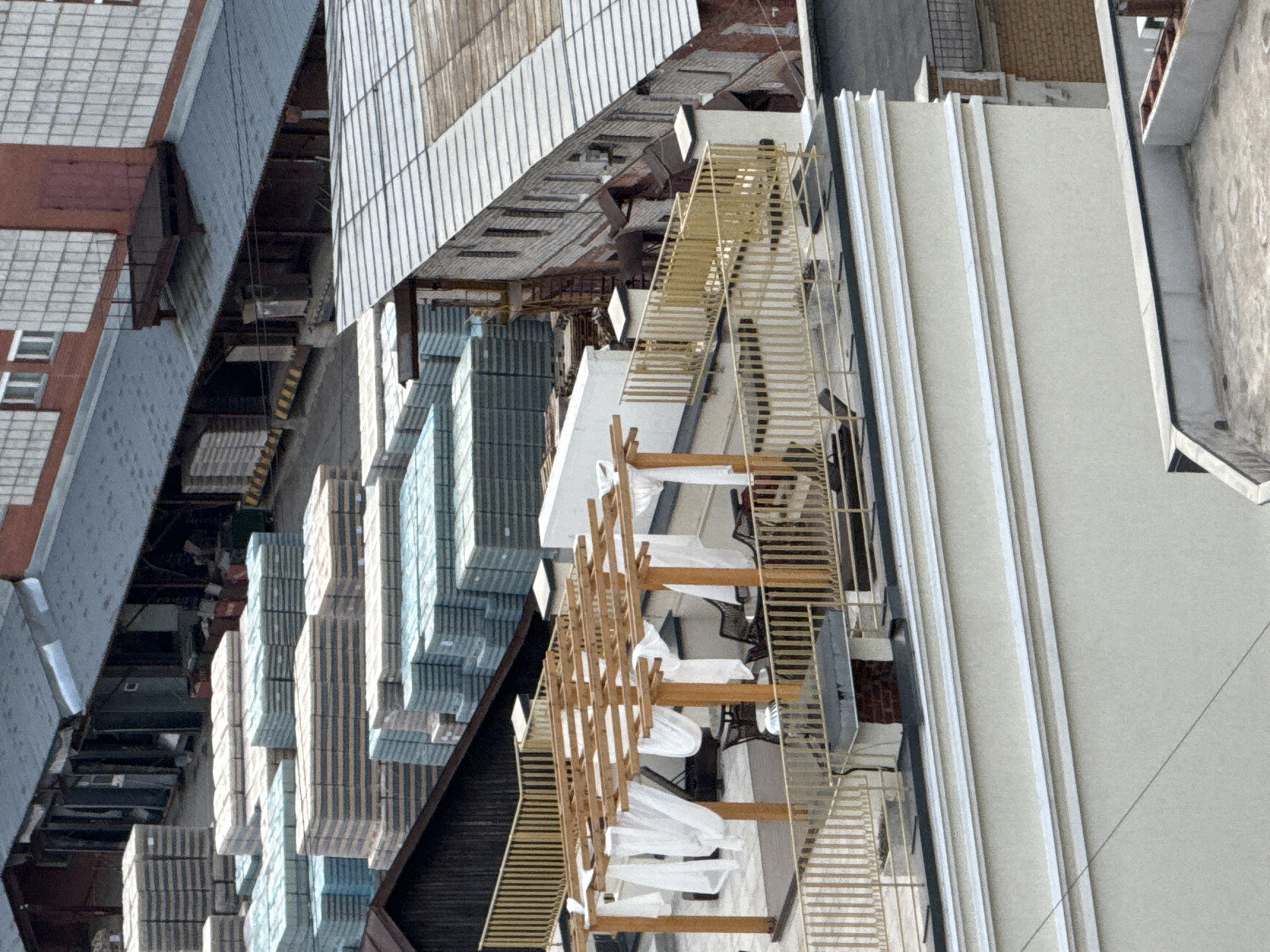 An aerial view of an industrial zone, potentially within the city of Perm, Russia, reveals stacks of building materials, possibly insulation or roofing, arranged in neat rows within a narrow alleyway formed by two large structures. Adjacent to the stacks is a wooden pergola-like structure covered with white fabric, suggesting a temporary outdoor space, possibly a restaurant or event venue. A metal staircase runs alongside one of the buildings. The buildings themselves are constructed from various materials, including concrete and metal sheeting, indicative of utilitarian industrial architecture.