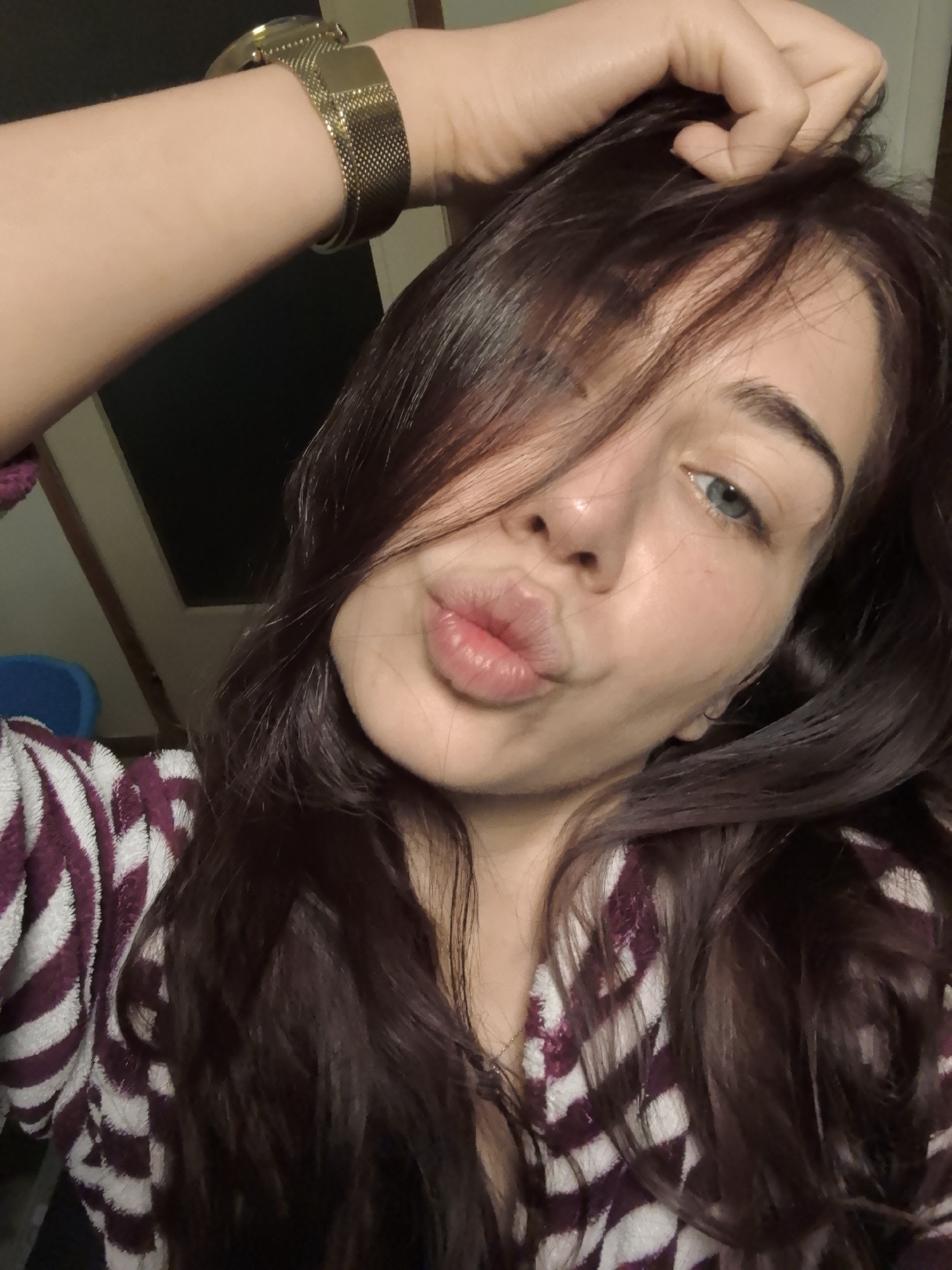 A young woman poses for a selfie. She has long, dark hair with some strands across her face, light skin, and blue eyes. She is puckering her lips slightly. She is wearing a purple and white striped robe and a gold watch. Her hand is raised and resting on her head. The background is dark, with a door partially visible. The scene appears to be indoors. There is no clear indication of the location being in Stara Zagora, Bulgaria.