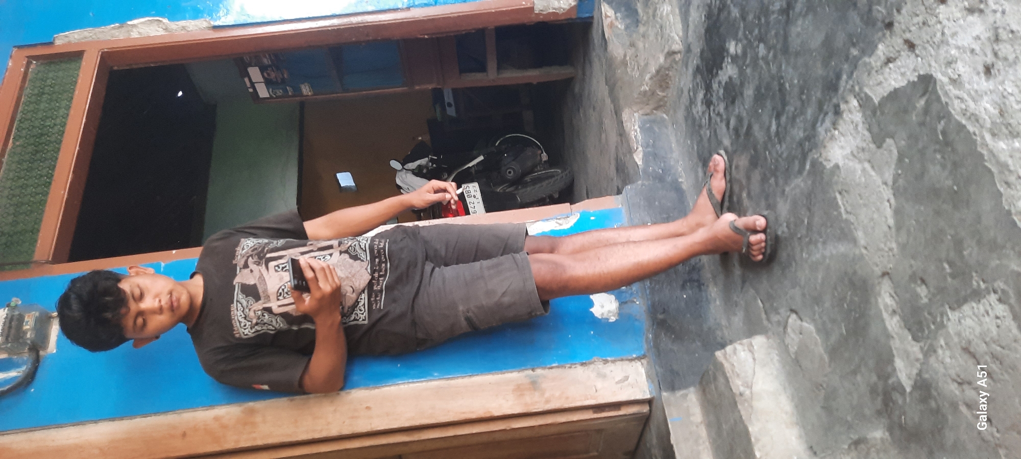 A young person, appearing to be of Indonesian ethnicity, stands leaning against a bright blue wall in what appears to be a residential area in Selopuro, Indonesia. The individual is wearing a dark t-shirt with a graphic print and dark shorts. They are holding a smartphone in their right hand, looking down at it. The person is wearing dark flip-flops. Behind them, an open doorway reveals a glimpse of an interior space with a brown door frame and a parked motorcycle with a license plate reading "642 DBS". The ground in front of the person is a weathered, grey concrete surface. The overall environment suggests an outdoor setting, possibly a front porch or entryway.