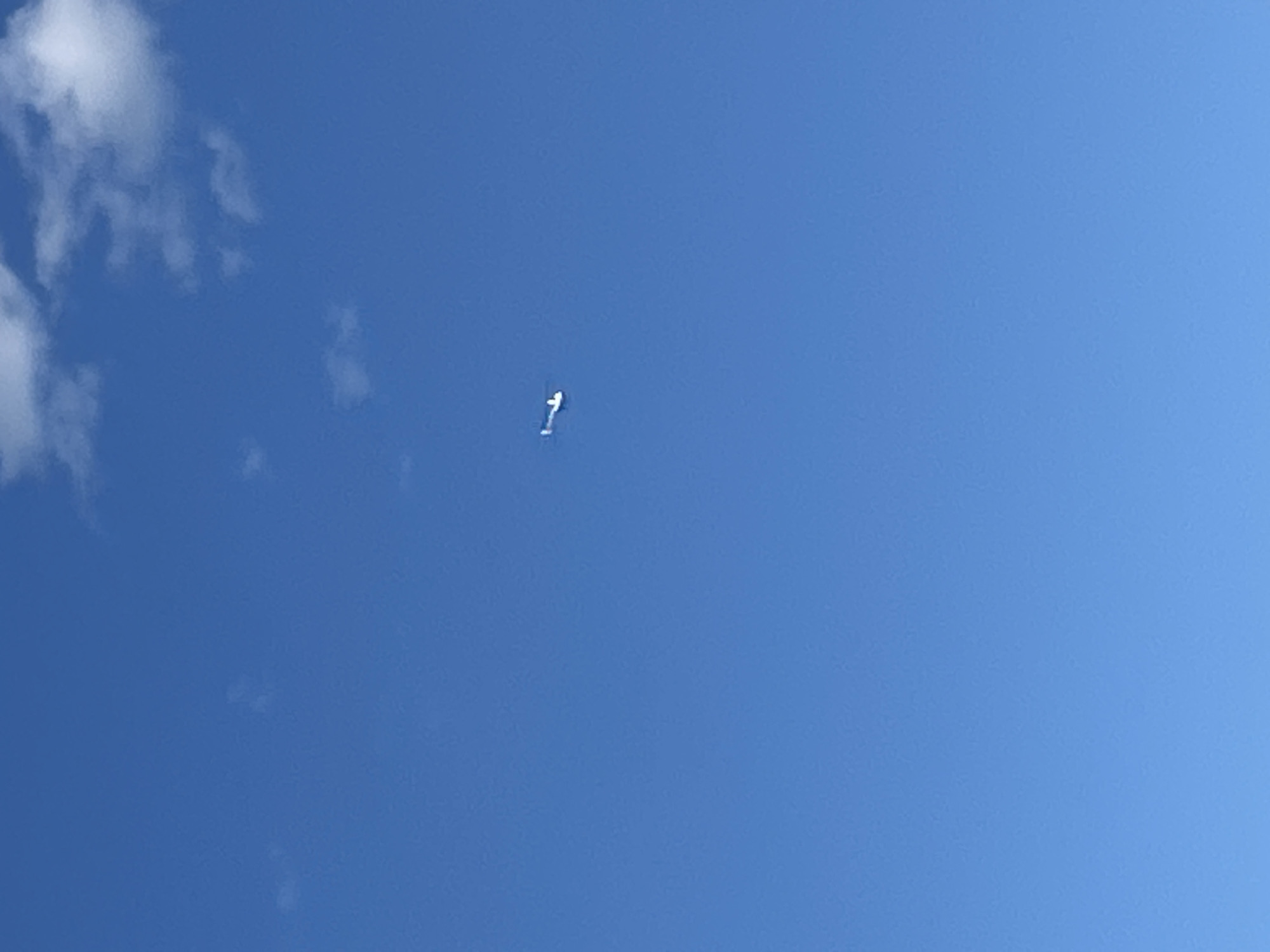 A white drone is captured mid-flight against a clear blue sky with a few white clouds. The drone is positioned in the center-right of the frame, appearing small due to its distance from the camera. The scene is an open sky, with no discernible city structures or land features visible. The location is specified as Слава Снт, Russia.
