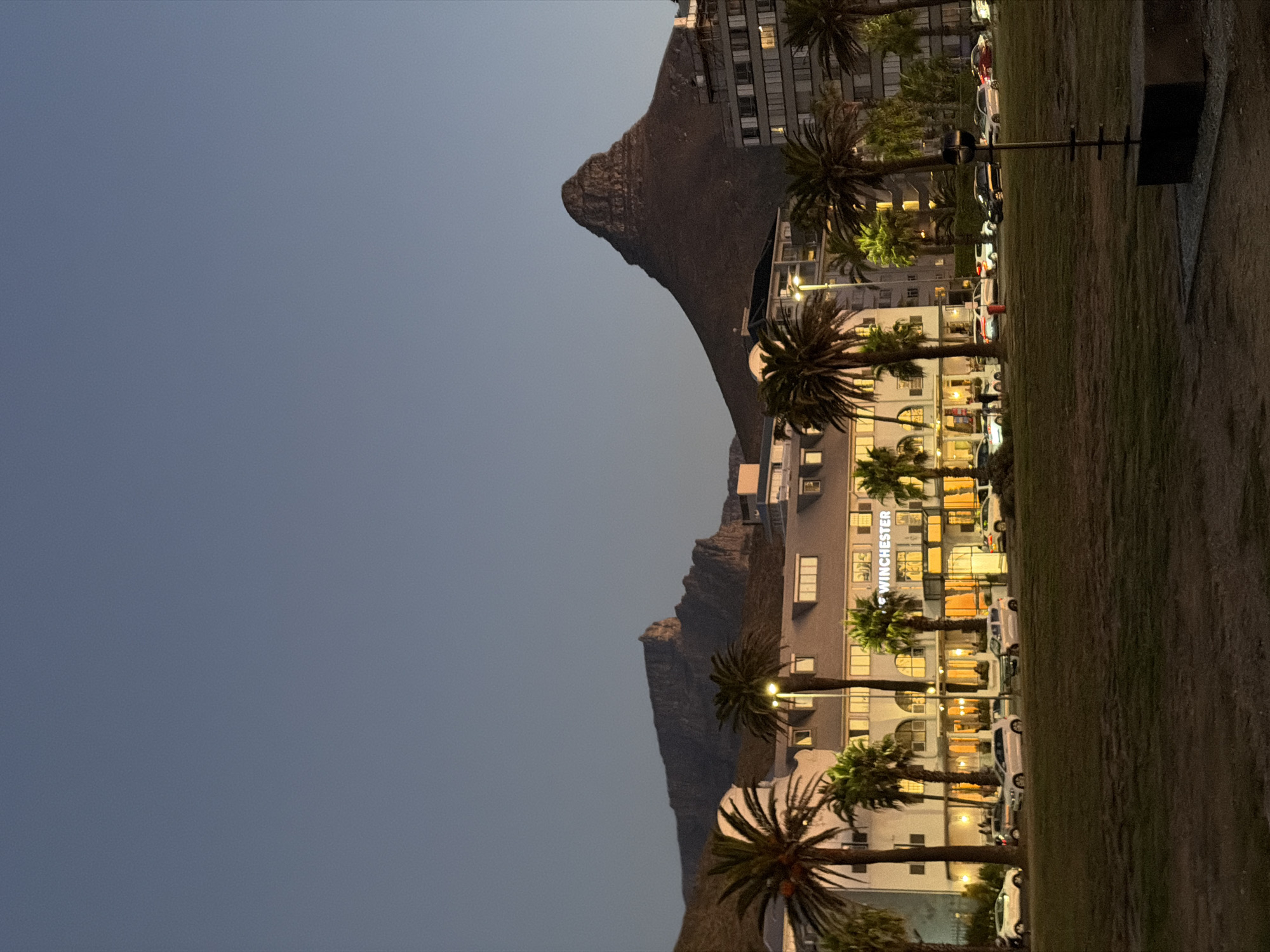 The image depicts a street scene in Cape Town, South Africa, at dusk or early evening. The sky is a gradient of blue. A mountain range, likely part of Table Mountain, is visible. Along the street, there are palm trees, buildings, and parked cars. One building, identifiable by the word "WINCHESTER," appears to be a hotel. A grassy area borders the street on one side. A metal object, possibly a light fixture, is also visible on the grass.
