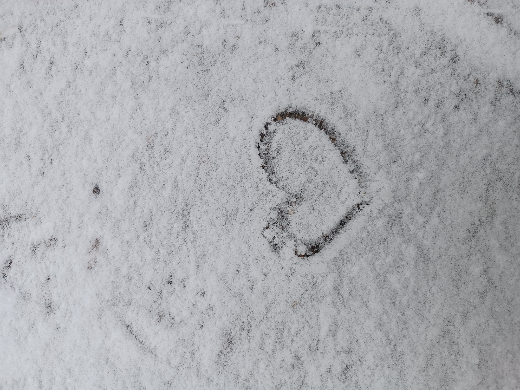 The image shows a ground surface covered in snow. A heart shape has been drawn in the snow, revealing the darker ground underneath. The location is Unknown, Unknown.