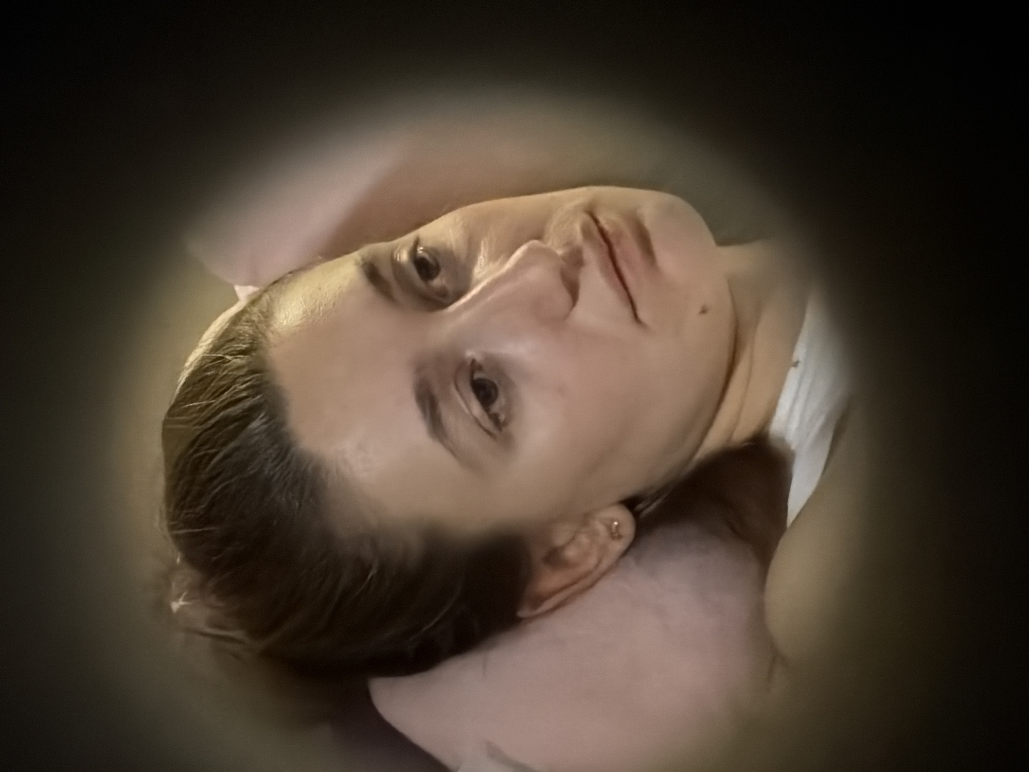 This portrait photograph features a light-skinned individual, likely female, positioned as if lying down, possibly in a bed. The subject has dark blonde or light brown hair and is resting their head on a light-colored, possibly pink, pillow. A white garment is visible on their right side, suggesting sleepwear or casual attire.
The person's face is the primary focus, illuminated against a dark, blurred background with a strong vignetting effect that emphasizes the central subject. They are looking slightly upwards and to the viewer's right with open, dark eyes, conveying a neutral to mildly pensive expression. The lips are slightly parted, and a small mole is visible on the upper lip. A small, gold-colored stud earring is visible in the right earlobe.
The lighting suggests a dimly lit indoor environment, potentially at nighttime. There are no specific weather or explicit location cues within the image itself, but the provided context places the location as Sofia, Bulgaria. No visible text is present in the media file. The overall atmosphere suggests a moment of quiet contemplation or rest.