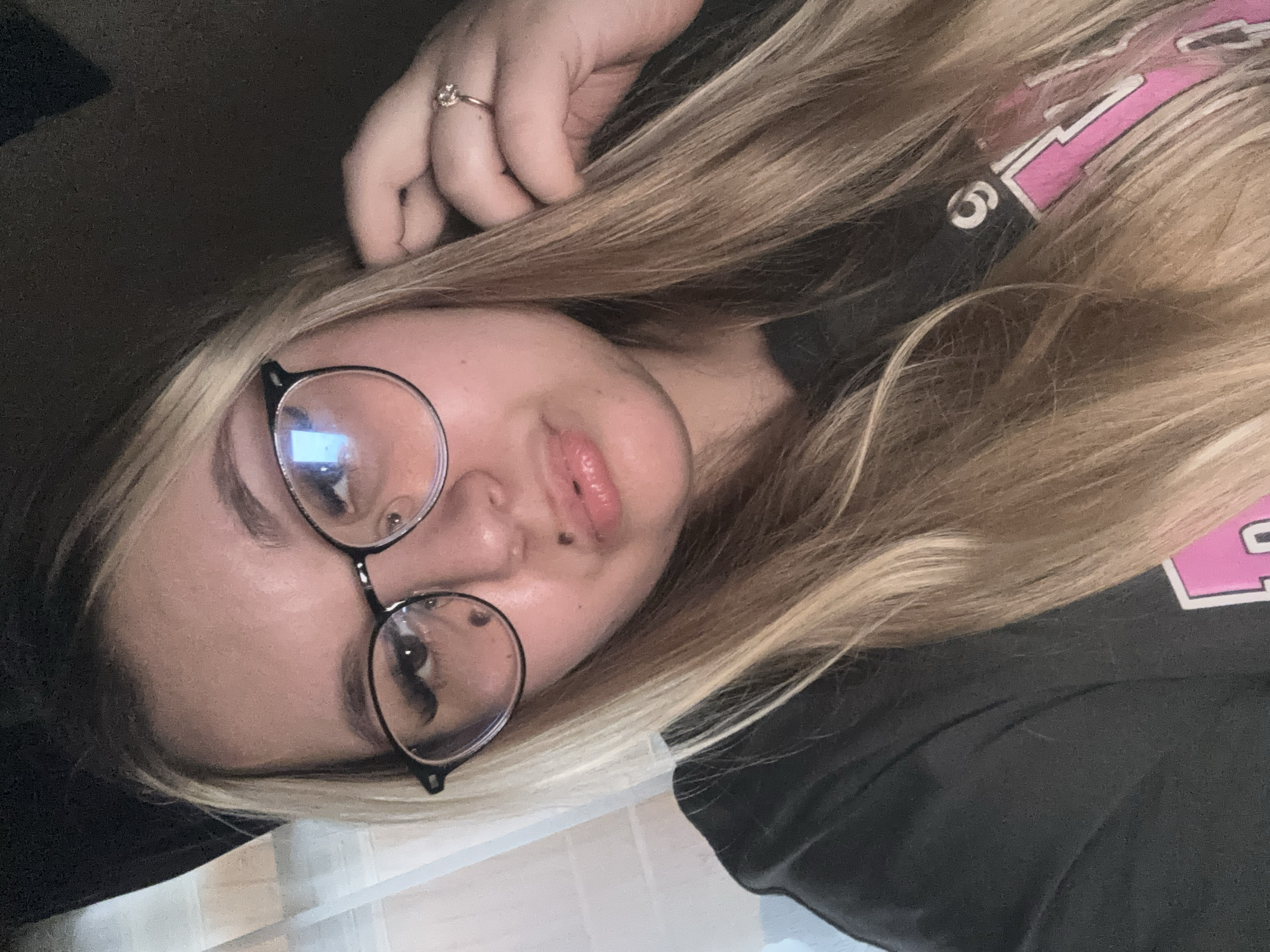 A young woman with blonde hair and glasses is posing for a selfie. She is wearing a dark t-shirt with pink lettering. Her left hand is touching her hair near her ear, and she is wearing a ring on her right ring finger. She has two beauty marks on her face, one on her cheek and one on her upper lip. The background is dark and out of focus. There is no indication of the location being in Bolshoy Bukor, Russia.