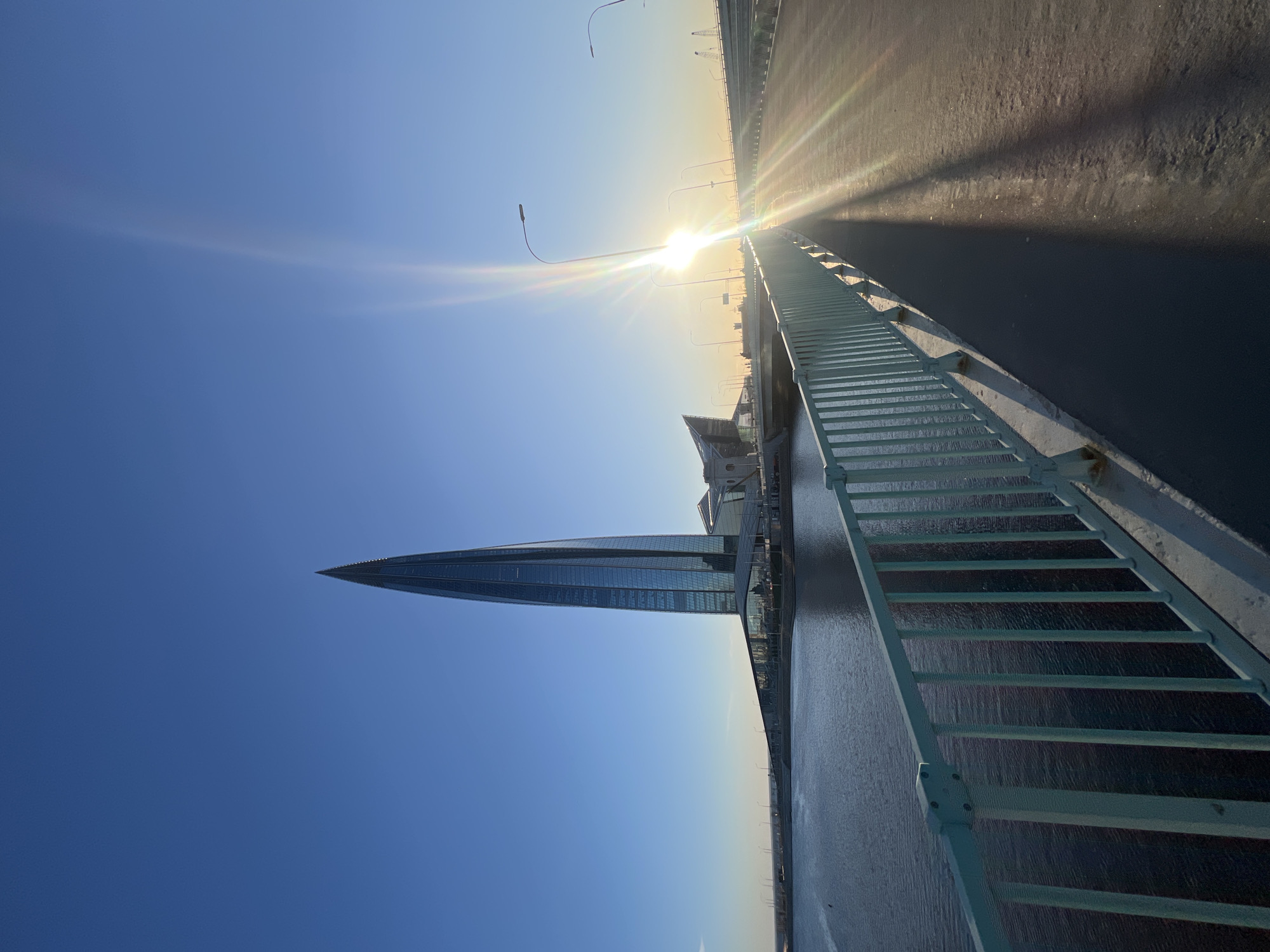 The image depicts a view from a bridge, possibly in Saint Petersburg, Russia, toward the Lakhta Center skyscraper. The sun is positioned on the horizon, casting a bright glare and long shadows on the road surface. A metal railing runs along the right side of the bridge. The sky is clear blue. The Lakhta Center is visible on the left, its pointed top reaching into the sky. The environment appears to be an urban setting with water visible below the bridge.