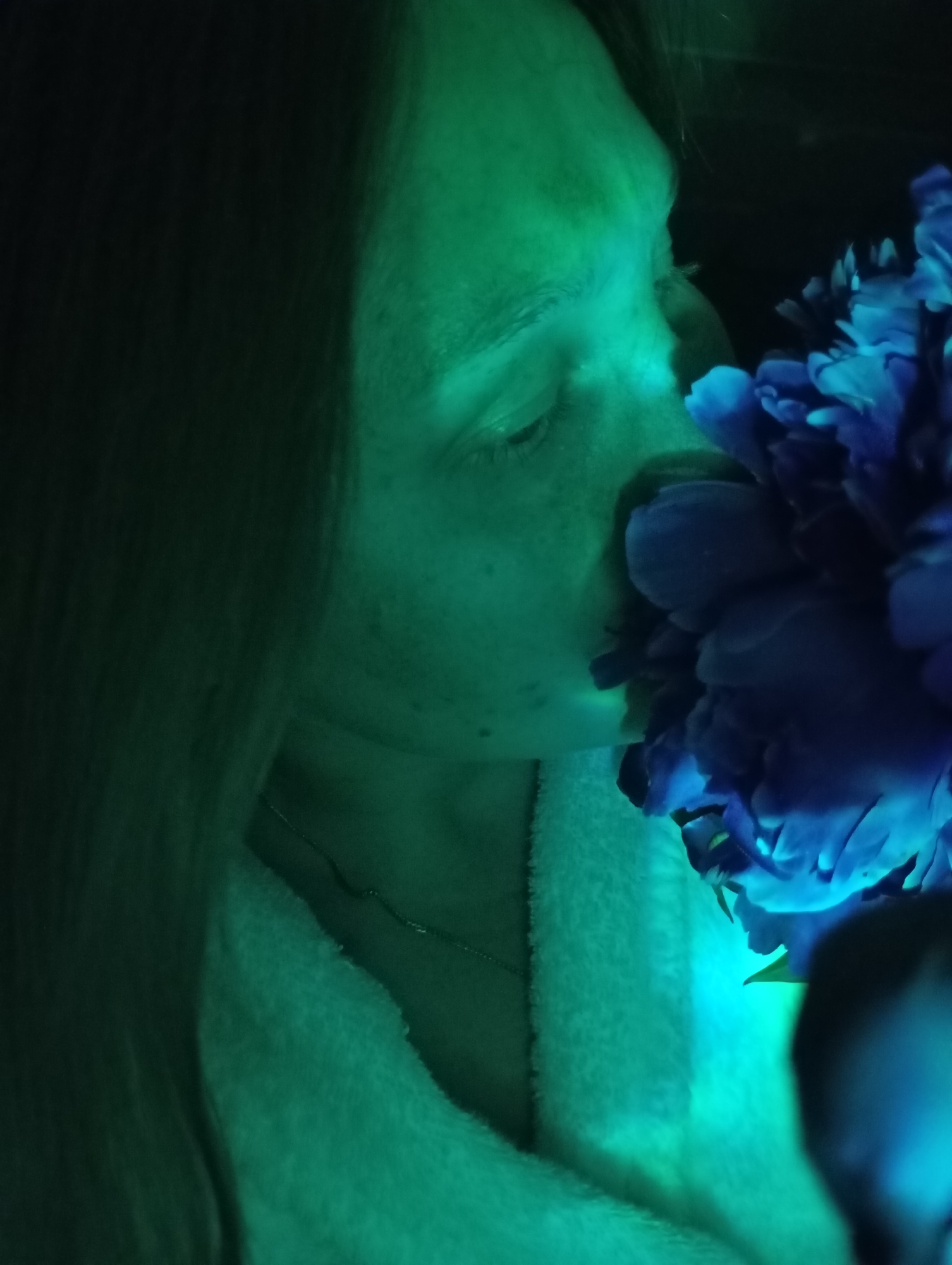 The image shows a close-up profile of a person's face, illuminated by a strong green light. The person is leaning in towards a vibrant blue flower, which is the central focus of the image. The person's eyes are closed, suggesting they are smelling the flower. A white, fluffy texture, possibly a robe or towel, is visible around the person's neck and chest. The background is dark, with some hints of hair or fabric. The overall mood is serene and intimate, with the artificial lighting creating a dramatic effect. There is no discernible information within the image that would indicate a specific location in the city of Fokino, Russia.