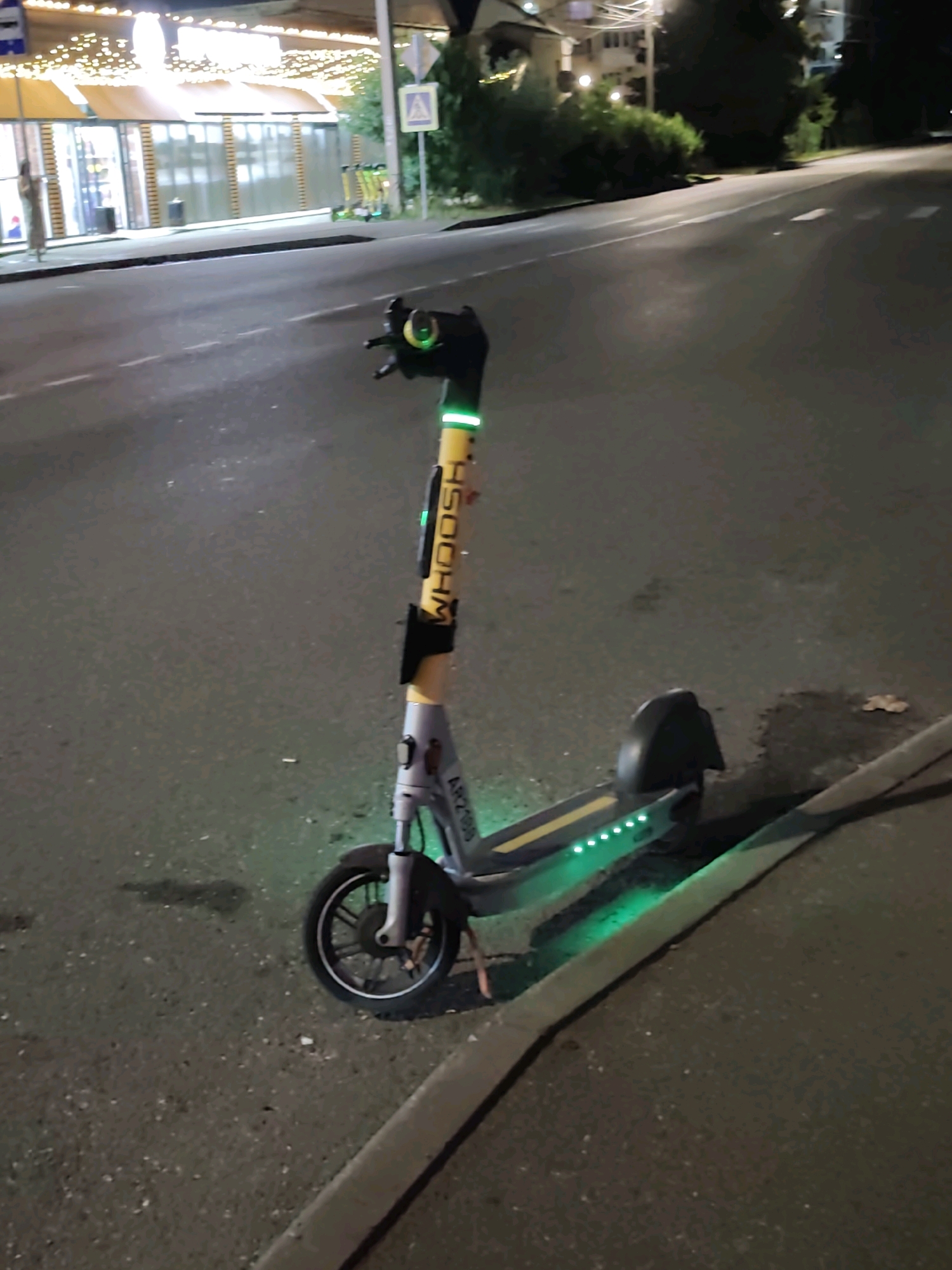 An electric scooter is parked on a sidewalk next to a road at night. The scooter is gray and yellow with green lights on its deck and handlebars. The word "WHOOSH" is visible on the scooter's yellow stem. In the background, there is a building with lights on and several other electric scooters parked in front of it. The scene appears to be in an urban environment, possibly in Sochi, Russia, given the style of the building and the presence of other scooters. The road has white lane markings, and there are trees and streetlights visible in the distance. The sidewalk is made of asphalt, and there is a curb separating it from the road.