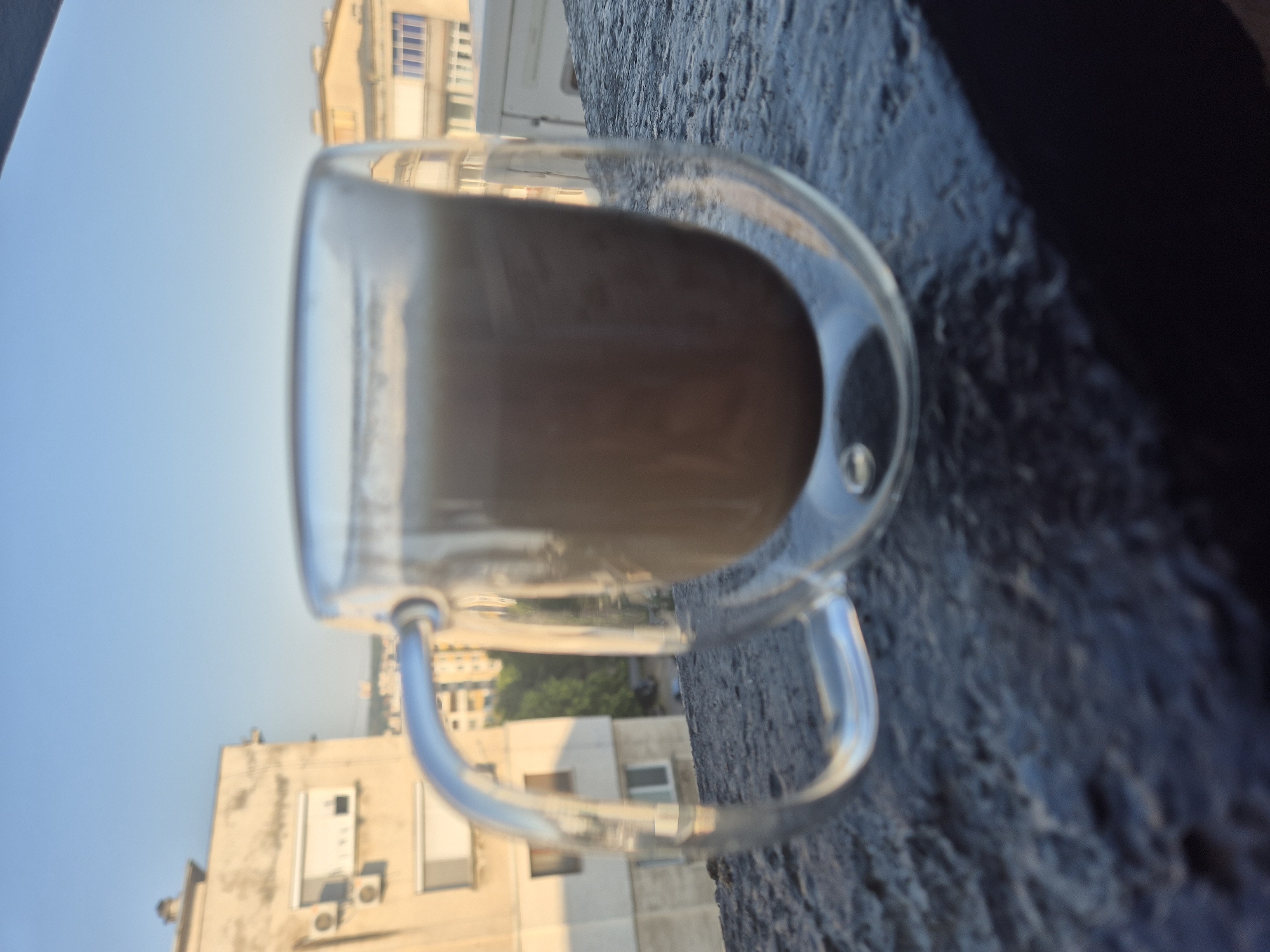 A clear glass mug containing a light brown beverage sits on a dark, textured surface. The mug has a double-walled design and a clear handle. In the background, a city skyline is visible, with buildings of varying heights and colors under a clear blue sky. The light suggests it is either early morning or late afternoon. There are no people or animals present in the image.