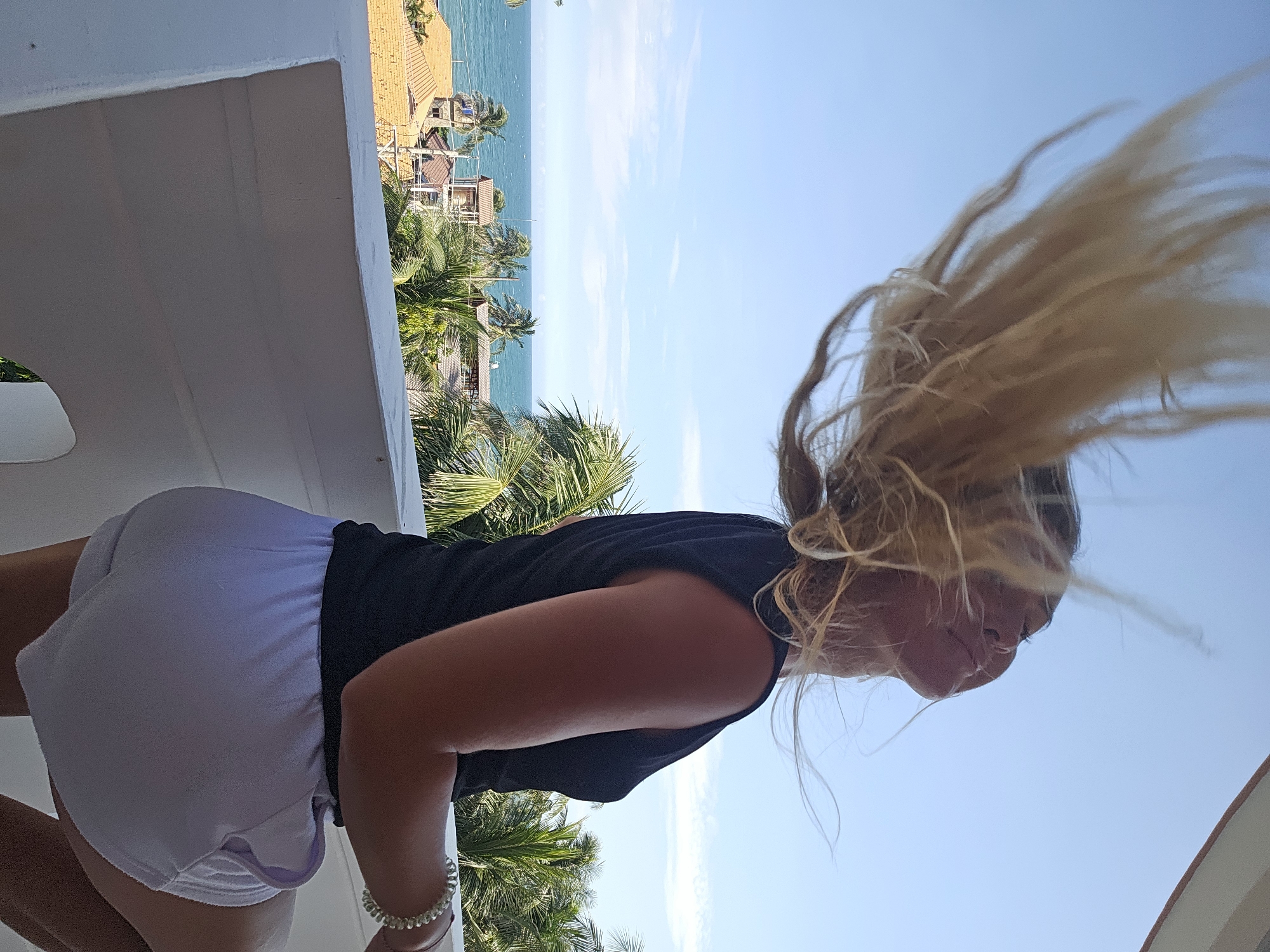 A blonde woman with her head tilted back and hair flowing in the wind stands on a white balcony. She wears a black sleeveless top and white shorts. In the background, palm trees and buildings are visible against the ocean and a clear blue sky. The location is identified as Phu Thuy Ward, Vietnam.