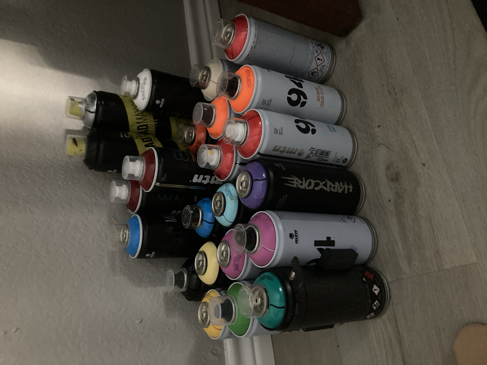 A collection of spray paint cans is arranged on a light-colored wooden floor against a neutral-colored wall. The cans are of various colors and brands, including MTN 94, MTN Hardcore, and MTN Water Based. Some cans are upright, while others are tilted or leaning. The lighting in the scene is dim, casting shadows on the wall and floor. There is no visible indication of the specific location within Round Rock, United States of America, but the environment suggests an indoor setting.