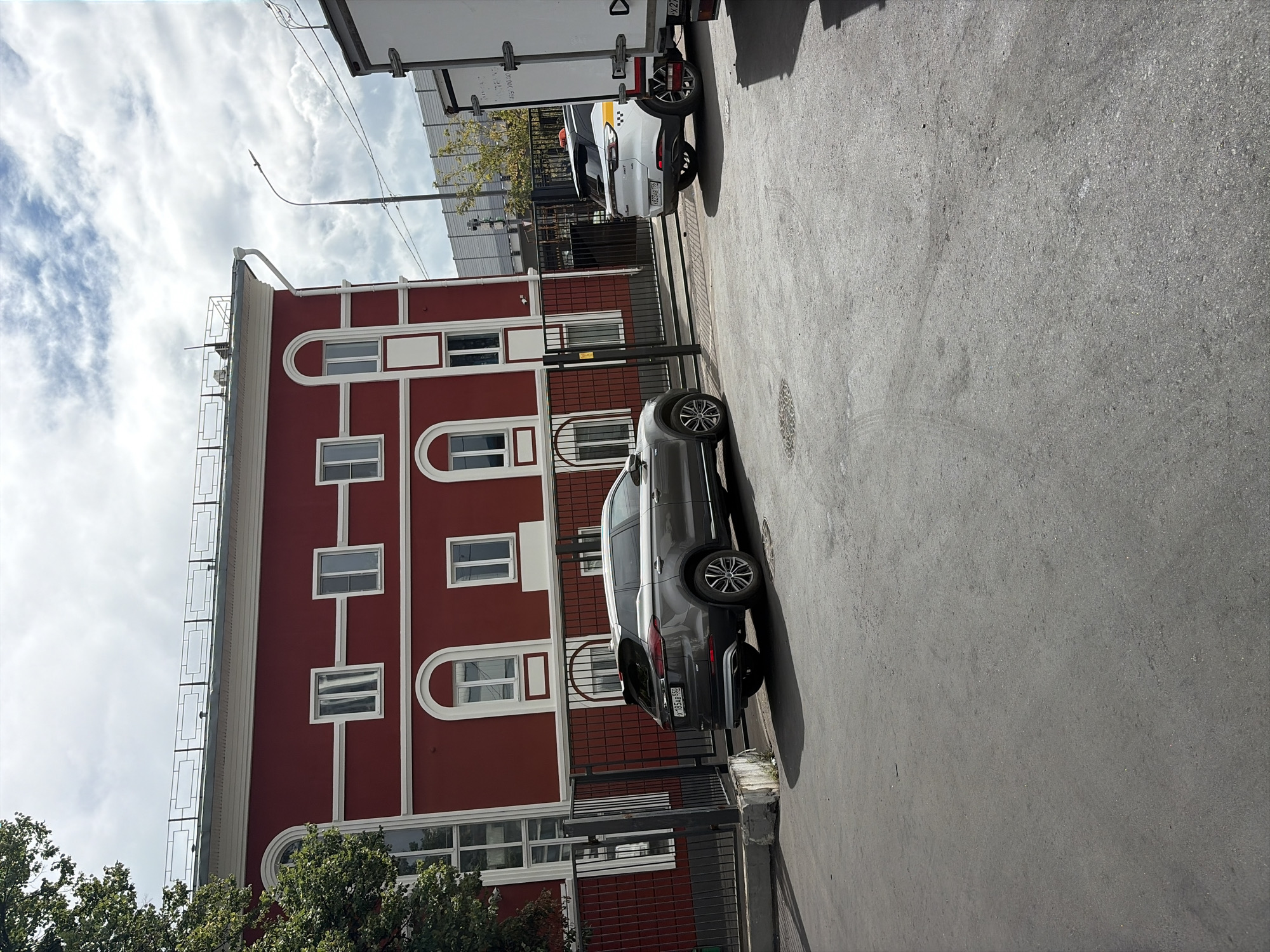 The image depicts an outdoor scene in Moscow, Russia. In the foreground, a gray SUV is parked on an asphalt surface. To the right of the SUV, a white car with a taxi marking is also parked. Behind the cars, a red building with white trim and arched windows is visible. A black metal fence runs along the front of the building. In the background, a white truck is partially visible on the right side of the frame, and a street lamp stands on the left. The sky is partly cloudy. There are no people visible in the image.