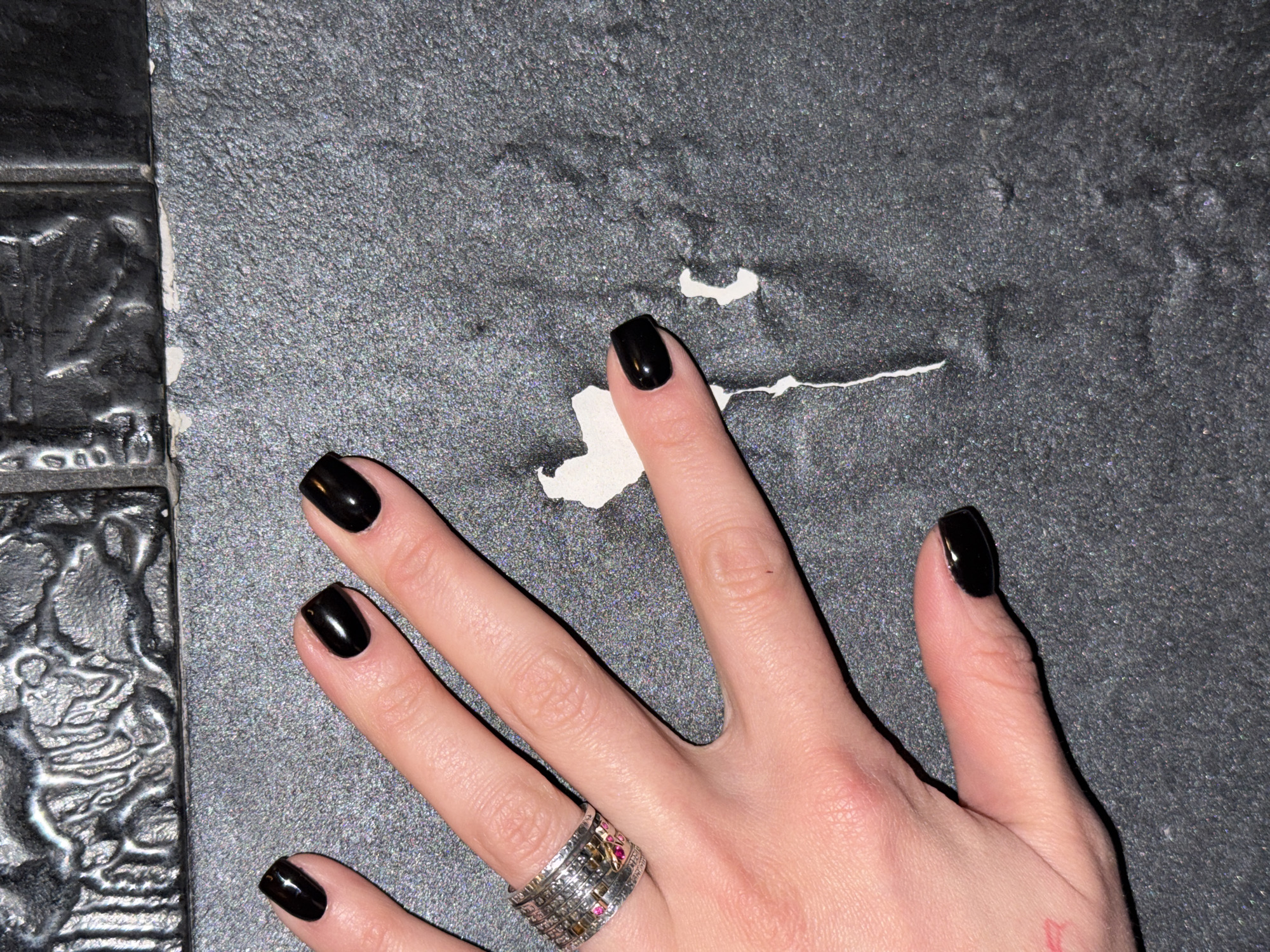 A right hand with polished black nails rests on a gray textured surface with a torn white section. The hand wears a wide, ornate silver ring with colored stones. There appears to be a faint red marking or tattoo on the lower part of the thumb. To the left of the hand, a decorative black tile is partially visible. The scene does not inherently depict or suggest the city of Sofia, Bulgaria, or any location. There are no interactions, actions, or additional subjects in the frame.