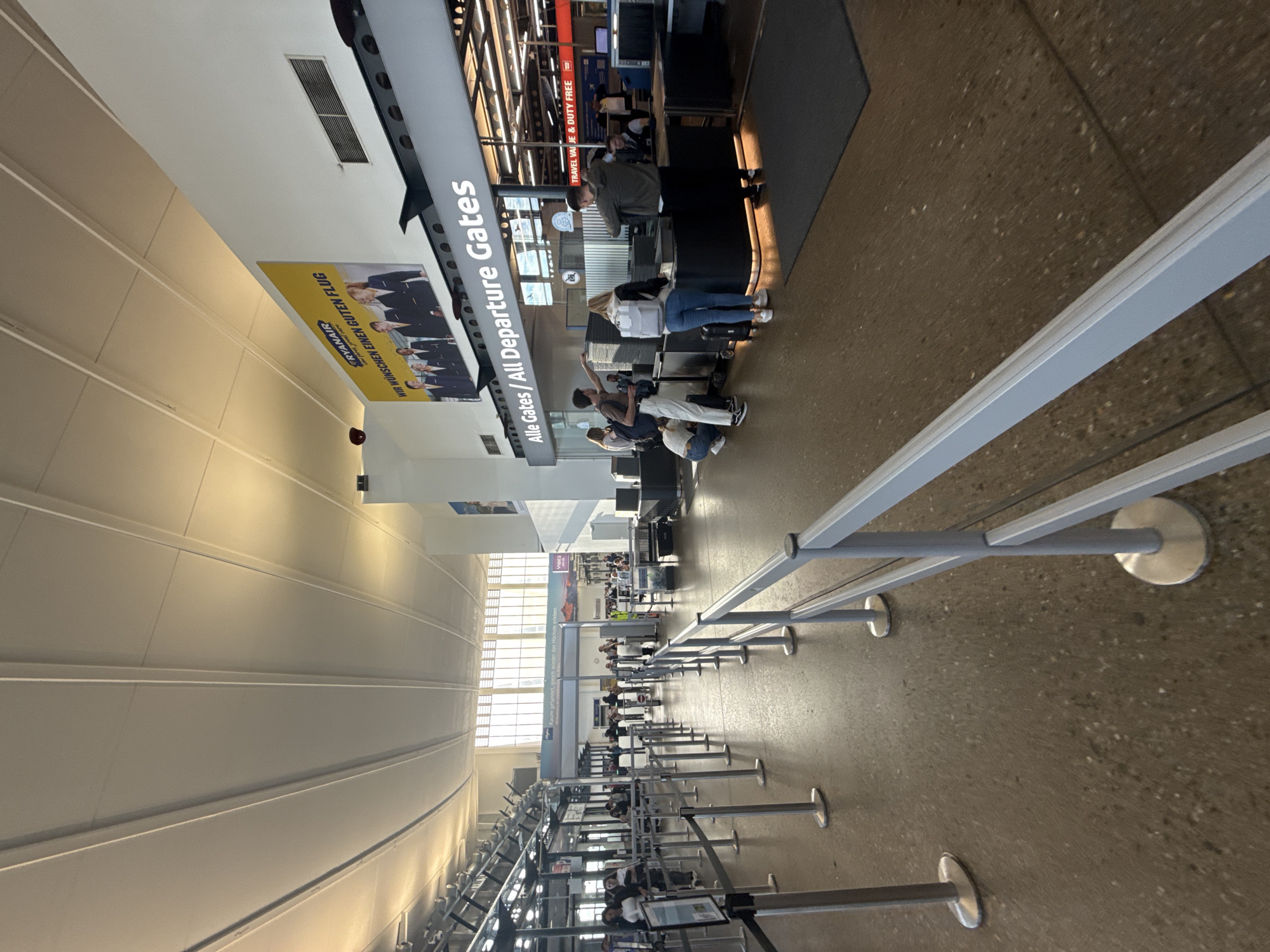 The image shows an interior view of an airport terminal in Künersberg, Germany. The scene depicts a security checkpoint area with queuing barriers and several individuals undergoing screening. To the right, a counter is set up with a scanner, and a person in uniform appears to be assisting passengers. Overhead, a large banner advertises Ryanair with the text "WIR WÜNSCHEN EINEN GUTEN FLUG" (We wish you a good flight). Above the checkpoint, signage reads "Alle Gates / All Departure Gates". The terminal features a high, curved ceiling with exposed lighting and ventilation systems, and the floor is a speckled terrazzo material. Natural light streams in from large windows in the background, illuminating the space. Several people are visible in the queues, some with luggage, and others interacting with security personnel.