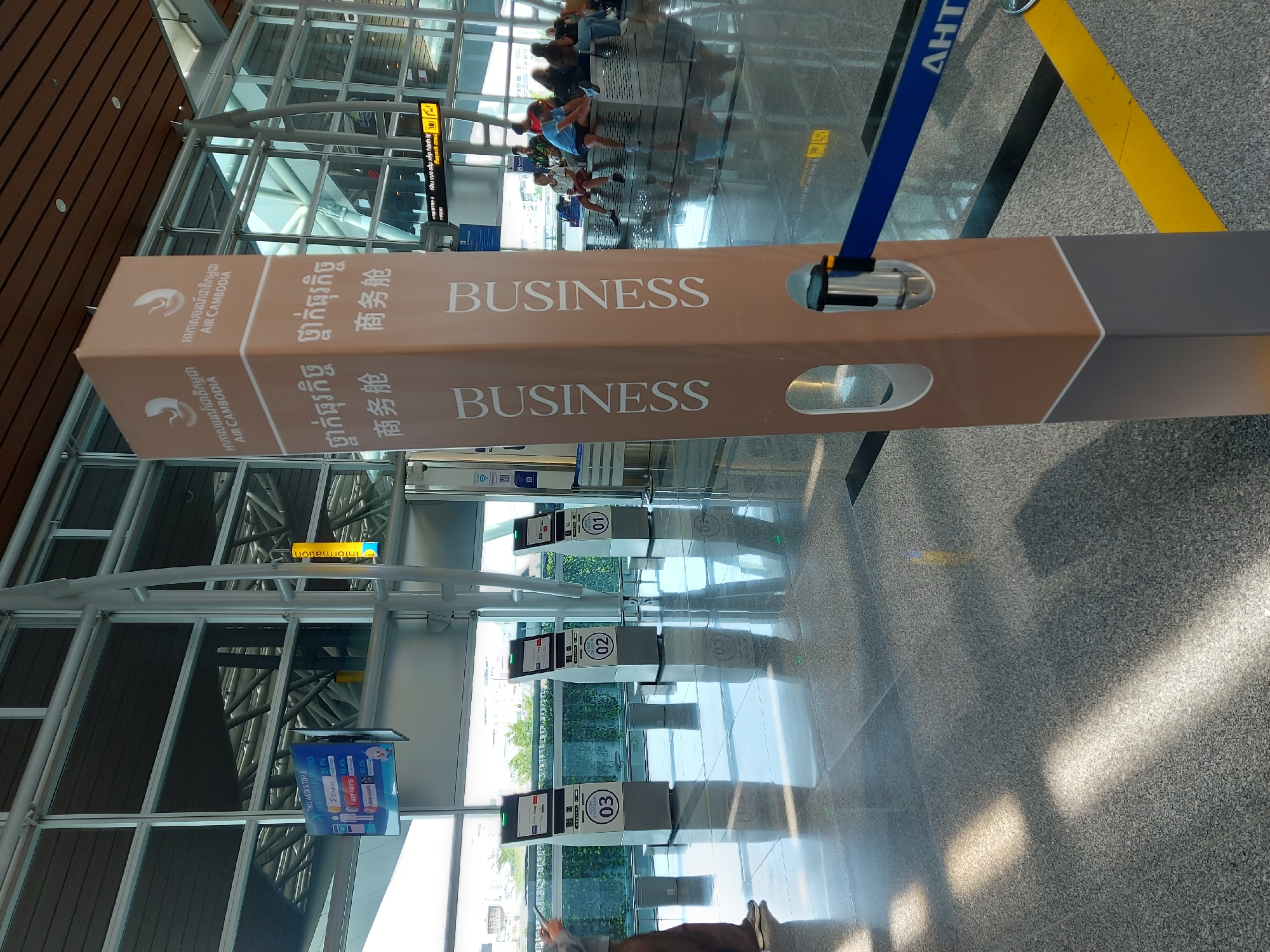 This image depicts the interior of an airport, likely in the city of Đà Nẵng, Vietnam, characterized by its modern architecture. In the foreground, a tall, beige-colored pillar stands prominently, featuring the "AIR CAMBODIA" logo and text in Khmer, Chinese, and English. The pillar also has the word "BUSINESS" printed vertically in English. A retractable blue belt barrier is attached to the pillar. To the left of the pillar are four self-service check-in kiosks labeled "03", "02", and "01". In the background, passengers are seated on benches, and a flight information display screen is visible. The floor is made of polished grey tiles, reflecting the ambient light. The overall environment suggests a functional and organized airport terminal.