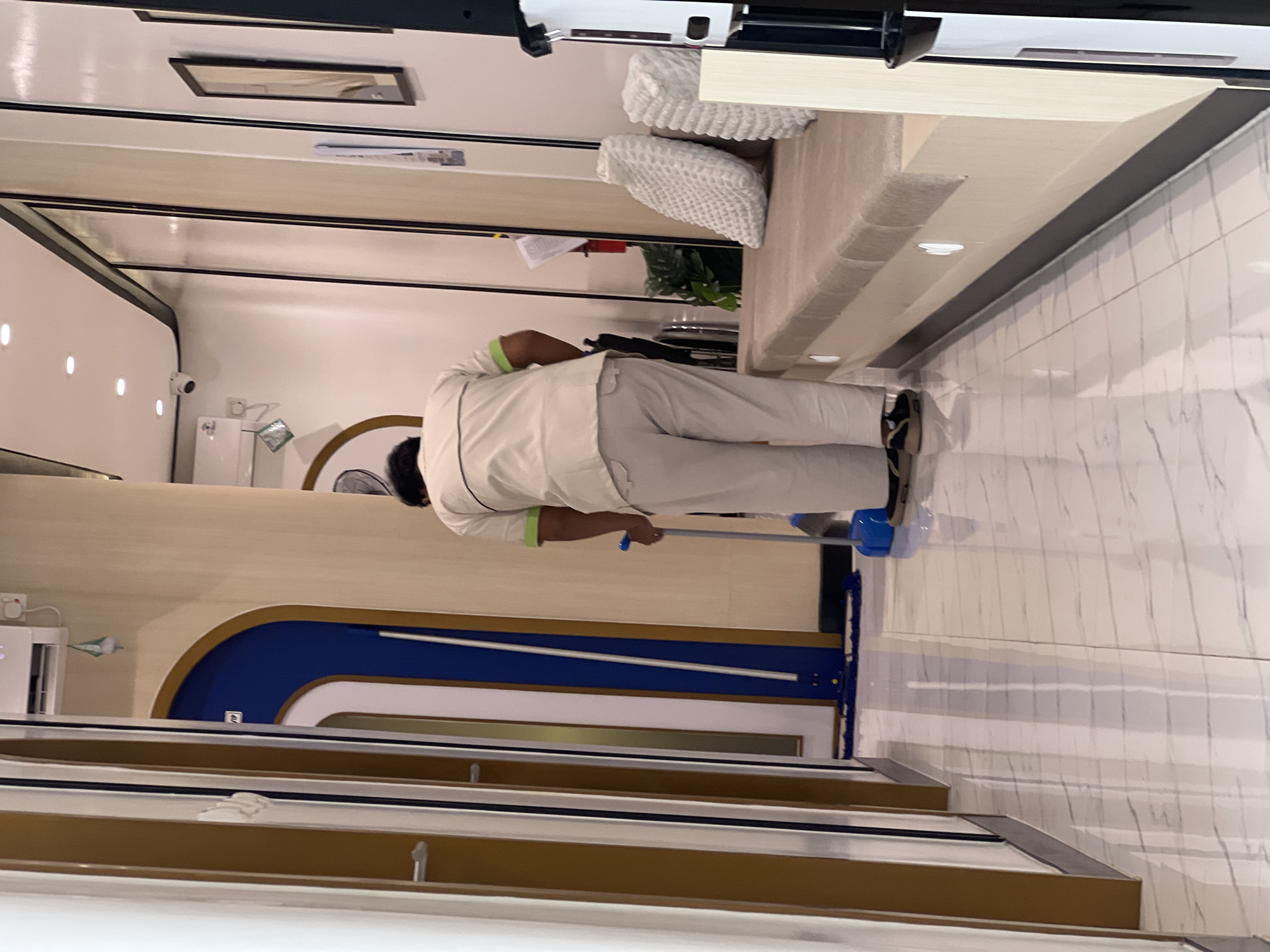 An unidentified individual, wearing a light-colored uniform with green accents, is sweeping a hallway with a blue mop. The hallway features polished marble floors, light-colored walls, and tall, slender doors with dark frames on the left. To the right, a cushioned bench is visible, with two textured white pillows and a large potted plant. In the background, a blue arched doorway with a white trim is present. The scene appears to be an interior location in Denpasar, Indonesia, likely a waiting area or a service establishment, given the presence of a bench and the clean, well-maintained environment.