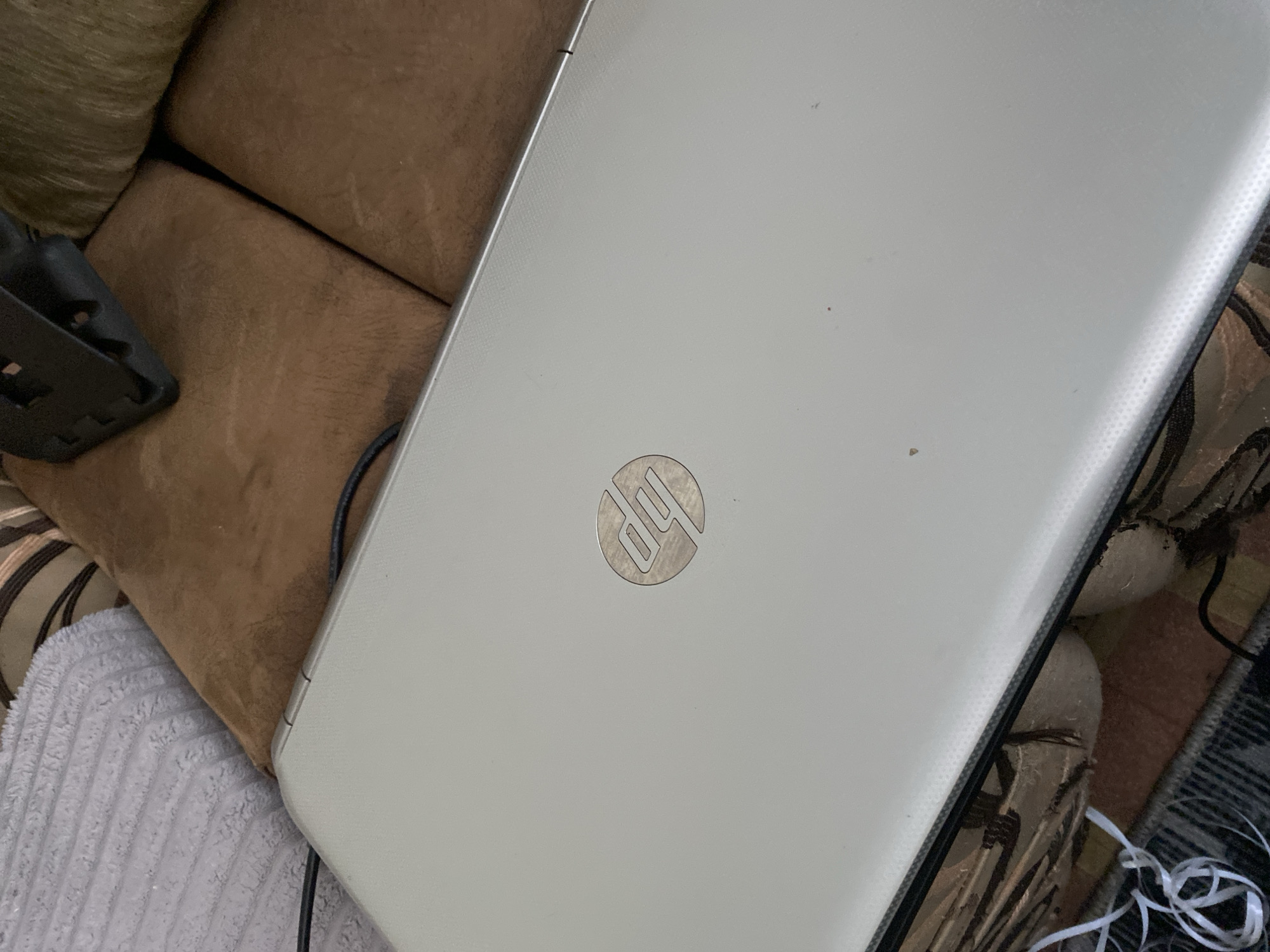 The image shows a closed, silver HP laptop lying on a brown upholstered surface, possibly a couch or armchair. The laptop lid features the distinctive metallic HP logo in the center. To the left of the laptop, a white cable is visible, partially uncoiled. Behind the laptop, on the right side of the frame, a black object, potentially a laptop stand or another electronic accessory, is partially visible. The background consists of the brown upholstery with a striped pattern and a textured gray material, likely a blanket or cushion, to the upper left. The scene is indoors. There is no information in the image that would allow for pinpointing a location in Chernogorsk, Russia.