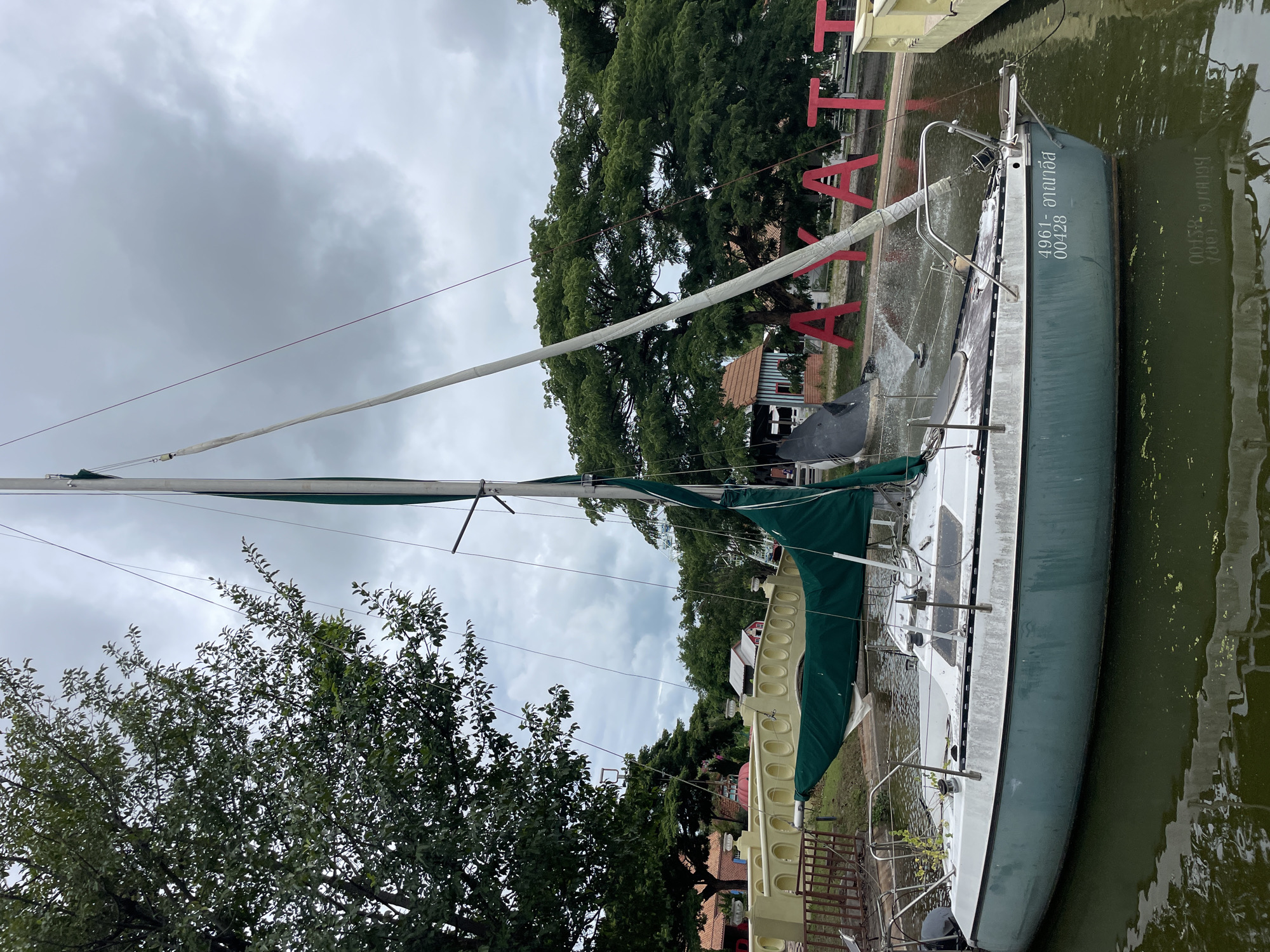 The image shows a white and green sailboat docked in a narrow waterway in Na Chom Thian, Thailand. The boat has a green sail furled around the boom. There is a number "4961- อาณาธิส 00428" written on the side of the boat. In the background, there are large trees, red letters spelling "AYATT" on the shore, and a yellow fence. The sky is overcast. The water appears murky with some floating debris.