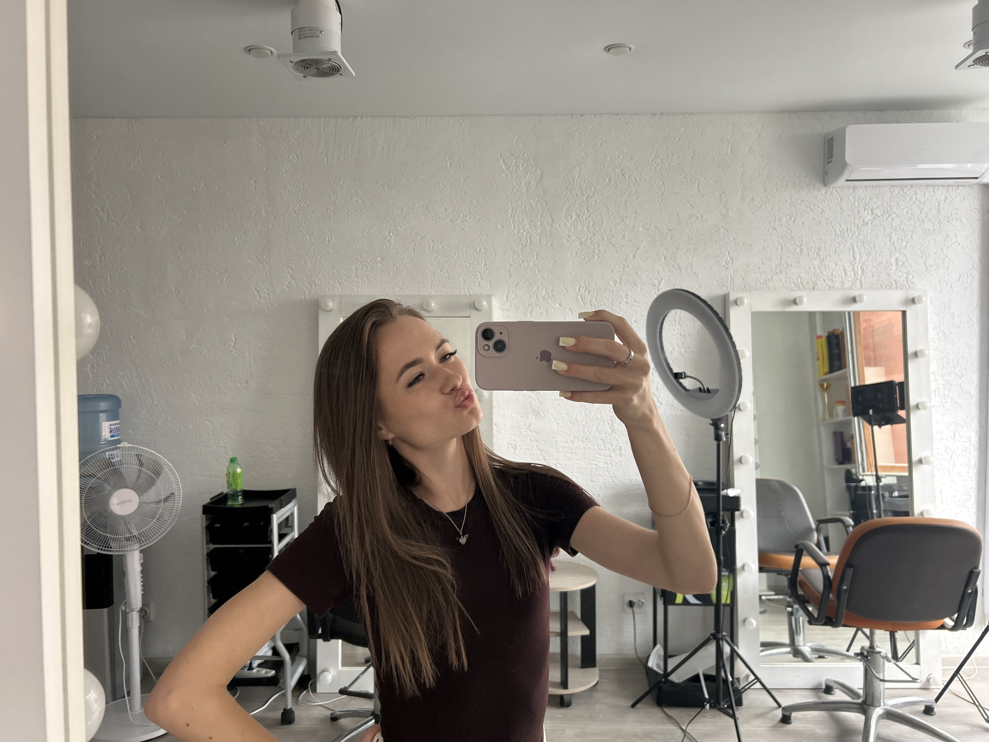 In an indoor setting, likely a beauty salon or dressing room, a young woman with long, brown hair takes a selfie in a mirror. She is wearing a dark burgundy t-shirt and a silver necklace. Her right arm is raised, holding a pink smartphone to capture the image. Her lips are pursed for a kiss. In the background, several items are visible: a large white fan, a water dispenser, a black trolley with shelves, a ring light on a tripod, and a white mirror with lights. The walls are textured and painted white, and an air conditioning unit is mounted high on the wall. The scene appears well-lit, with the natural light coming from an unseen source, possibly a window.