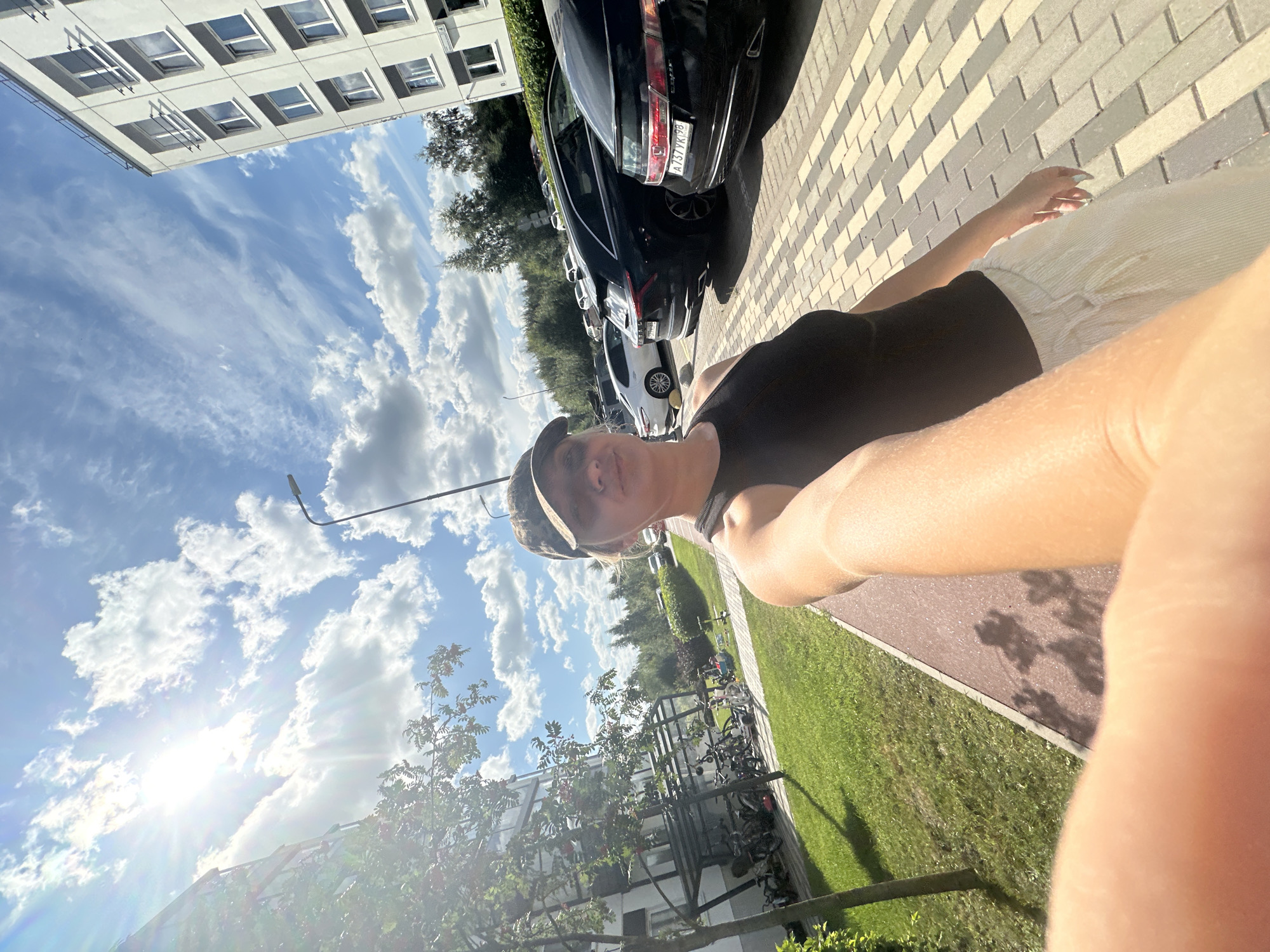 A young woman wearing a black tank top and a camouflage baseball cap takes a selfie in a parking lot in Vsevolozhsk, Russia. She is standing on a paved walkway with a row of parked cars to her right and a modern apartment building to her left. Sunlight streams through a partly cloudy sky, casting shadows on the ground. The parking lot appears to be well-maintained with a neat row of cars, including a black SUV and a black sedan with a license plate visible. To the left of the woman, there is a grassy area with trees and a bicycle rack with several bicycles. The woman is holding the camera in her right hand, and her left arm is bent at the elbow. The background shows more trees and buildings, suggesting an urban or suburban environment.