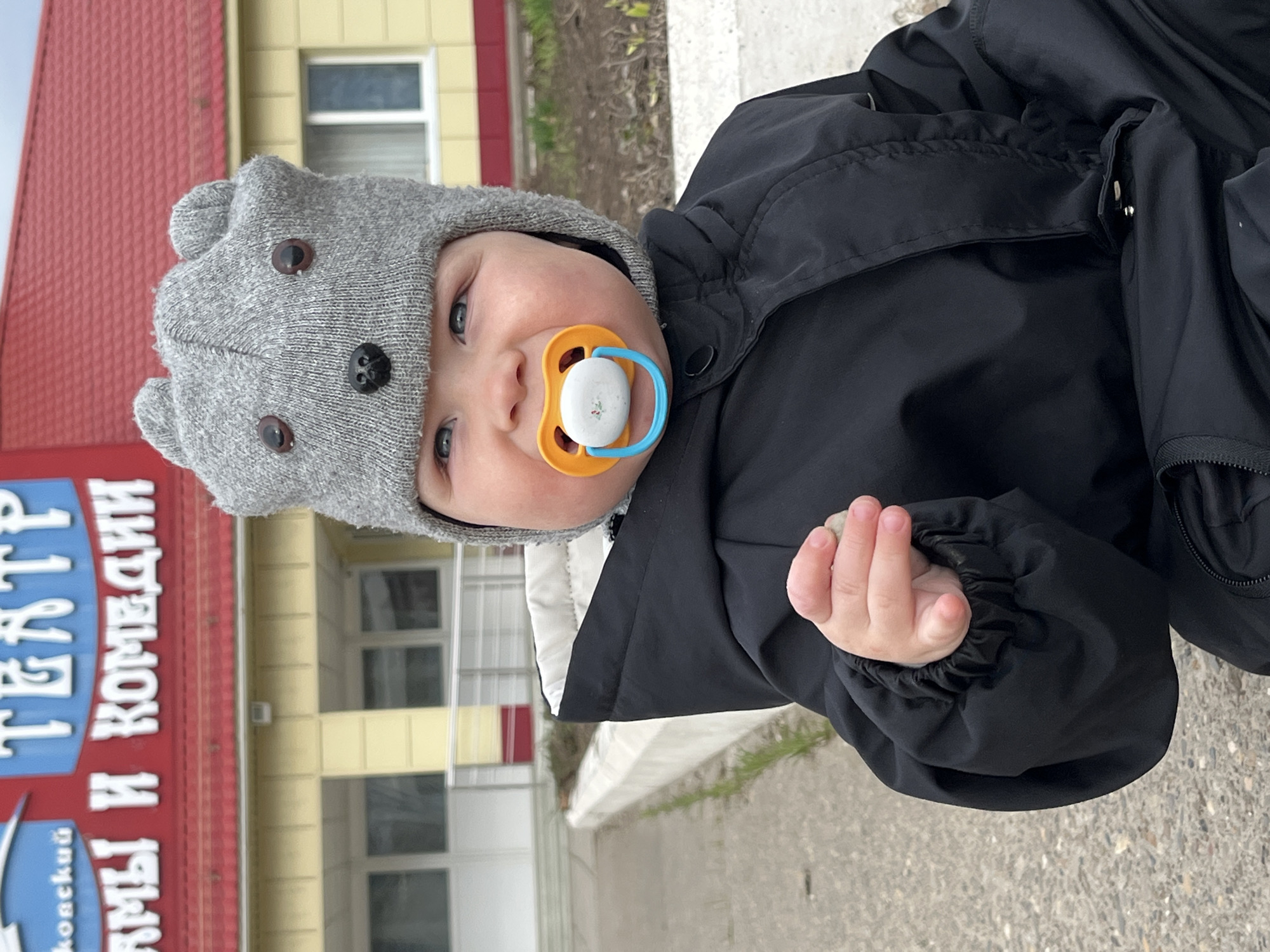 A baby wearing a gray hat with buttons resembling eyes and a black winter coat is holding a small rock in their right hand and has a pacifier in their mouth. They are standing on a concrete surface near a curb and some greenery. In the background, there is a building with a red roof and yellow walls, with white windows and a red sign that reads "Театр драмы и комедии" which translates to "Drama and Comedy Theater". The location is likely in the city of Chaikovsky, Russia.