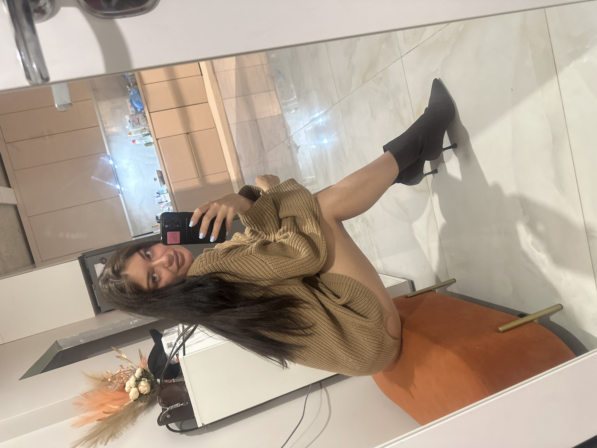 A woman in a brown sweater is reflected in a mirror while sitting on an orange chair, holding a cell phone. The reflection shows a beige wall with cabinets and tiled flooring. The setting appears to be indoors, potentially a residence. The image has been rotated; the location of Khimki, Russia is not confirmed from the image.