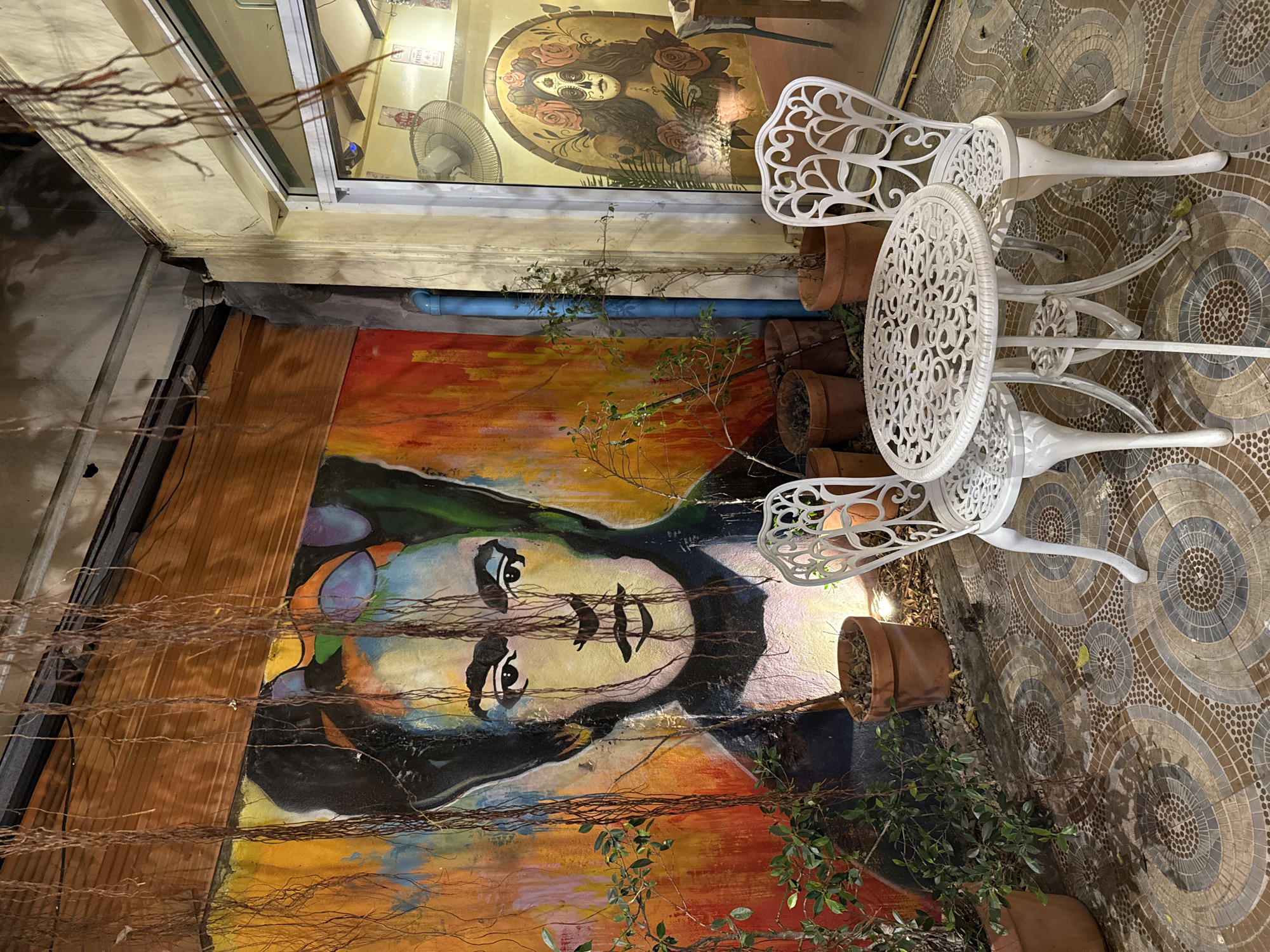 This image displays an artistic outdoor patio or courtyard scene at night in Baan Lamai, Thailand. A vibrant, colorful mural of a woman's face, reminiscent of Frida Kahlo with dark hair, strong eyebrows, and a contemplative expression, covers a large section of a vertical wall. The mural's background features a lively blend of red, orange, yellow, and blue brushstrokes.
In the foreground, numerous thin, dry, reddish-brown vines or branches hang vertically, partially obscuring parts of the mural and a wooden slatted structure to the left. On the ground, a decorative tiled floor, composed of circular and semi-circular mosaic-like patterns in shades of brown, beige, and grey, extends from the left to the right.
Arranged on this tiled floor are two white, ornate cast-iron chairs and a matching circular table, all adorned with intricate scrollwork and floral patterns. Several terracotta pots containing small plants or shrubs are placed around the base of the mural and near the furniture, some with sparse foliage, others with greener leaves.
Above the mural, a window with a white frame reflects interior elements, including a ceiling fan and another framed artwork, possibly a portrait. A light blue pipe runs horizontally along the wall between the window and the mural. The artificial lighting indicates it is nighttime or evening, highlighting the artistic details and creating a warm, inviting atmosphere. No specific activity is taking place, but the setup suggests a space for dining or relaxation. The word "FAN" is visible in the reflection within the window.