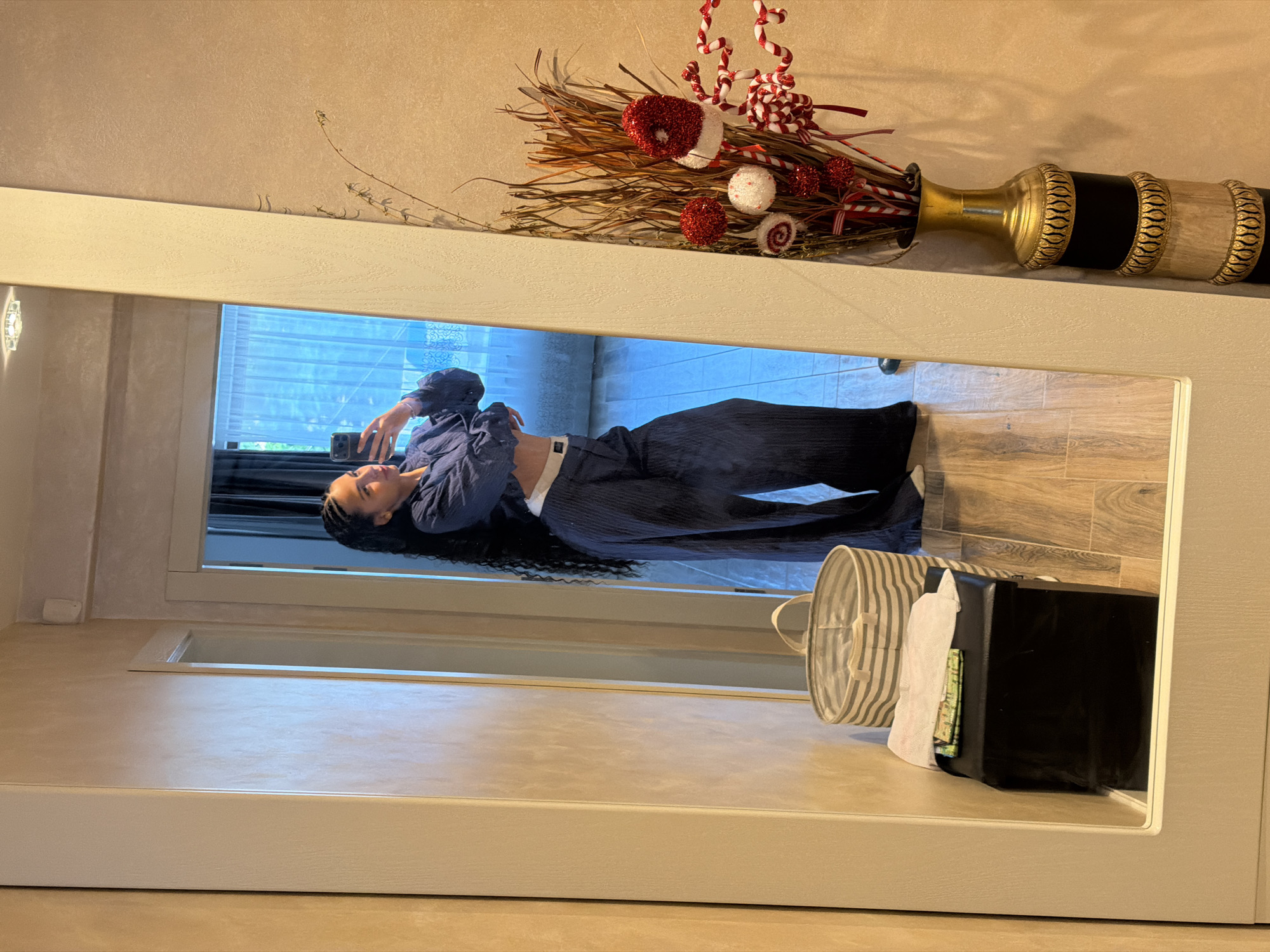 A full-body mirror reflection shows a young woman with long, dark, braided hair posing in a dimly lit interior space. She is wearing a dark blue collared shirt, unbuttoned at the bottom to reveal a white band, and matching dark blue wide-leg pleated pants. She is holding a smartphone in her right hand, angled to capture her reflection. The background visible in the mirror includes a doorway leading to a room with sheer blue curtains, sunlight filtering through, and a glimpse of greenery outside.
To the left of the reflection, a grey and white striped laundry hamper stands next to a black ottoman with a white cloth and a green item on top. To the right of the mirror, a tall, ornate vase filled with dried branches and festive red and white ornaments, including candy cane shapes and glittery red balls, is positioned against a textured beige wall. The floor appears to be made of wood-look tiles. The overall scene suggests a personal living space, possibly a hallway or dressing area, within a residence. The image is a candid, unposed shot taken by the individual. The location is not identifiable as Gabrovo, Bulgaria from the visual information provided.