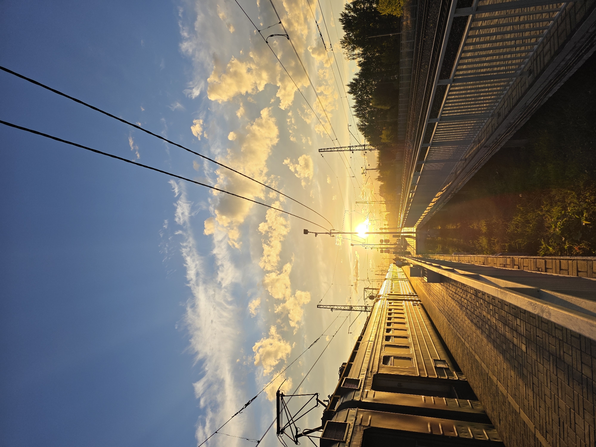 This image depicts a train station platform at sunset in Posolok Kombinata Stroymaterialov-2, Russia. A long train, partially illuminated by the setting sun, is parked at the platform. The platform itself is paved with brickwork, and a white railing runs along the edge, separating it from the train tracks. In the background, trees line the horizon, and overhead, power lines crisscross the sky, which is a blend of blue and warm sunset colors with scattered clouds. The sun, low on the horizon, casts long shadows across the platform. Two figures can be seen walking on the platform in the distance, silhouetted against the bright sky. The overall atmosphere is peaceful and serene, capturing a moment of quiet transition at the end of the day.