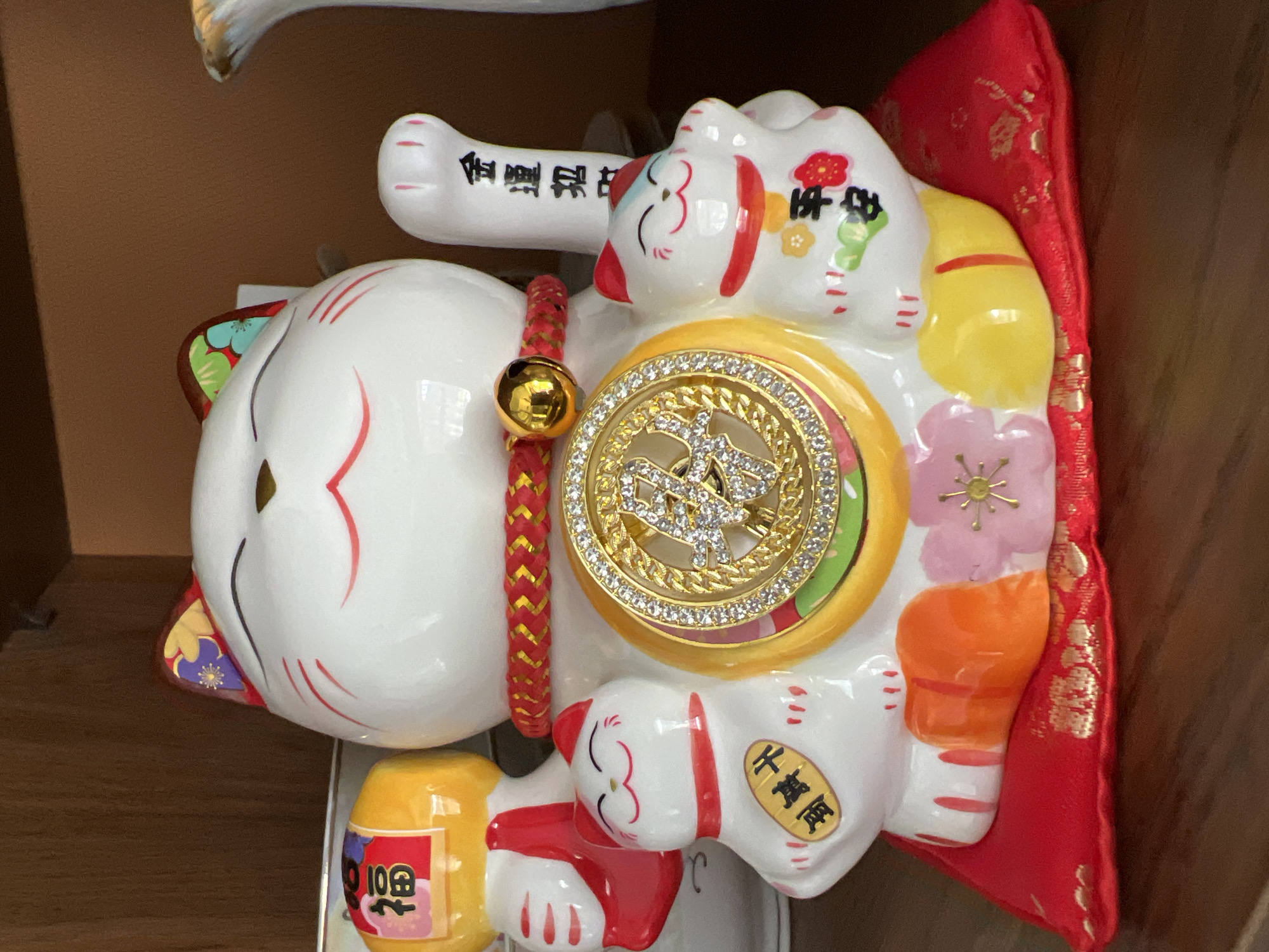 A ceramic Maneki-neko, or fortune cat, is displayed prominently on a red cushion, which rests on a wooden surface. The Maneki-neko is white with red markings on its face and ears. It wears a gold and red braided collar with a small gold bell. A circular, jeweled emblem adorns its chest, featuring gold characters. Two smaller cat figures flank the main one, one on each side, each with a red collar and a gold oval emblem on their chest with black characters. The main cat holds a yellow object with red writing in its left hand, and its right paw is raised in a beckoning gesture, with black characters inscribed on it. In the background, a wooden shelf is visible. The scene is indoors, and there is no information to suggest the location is in Еркін, Kazakhstan.
