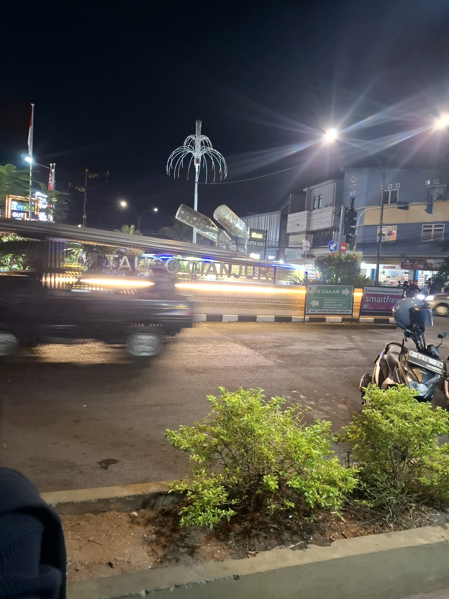 The image captures a night scene in Cianjur, Indonesia, likely an intersection with traffic. In the foreground, the edge of a concrete sidewalk is visible with lush green bushes planted along it. To the right, a black and gold motorcycle is parked, its license plate reading "F 6476 ZN". The road surface shows signs of wetness, suggesting recent rain.
Further back, a vehicle, possibly a public transport van or a small bus, is captured with motion blur, its headlights and taillights streaking across the frame. The vehicle has the words "TAIOGUATIAN" illuminated on its side, with bright yellow lights running along its top and bottom edges.
Across the street, buildings are illuminated, with signs indicating commercial establishments. One prominent sign reads "SMARTFREN". Another sign with arrows points to "SATLANTAS POLRES CIANJUR".
In the background, a large, abstract metal sculpture resembling a fountain or a futuristic tree stands illuminated against the night sky. A flagpole with the Indonesian flag (red and white) is also visible. Streetlights cast a bright glow, creating lens flares and illuminating the scene. The overall impression is that of a busy urban environment at night.