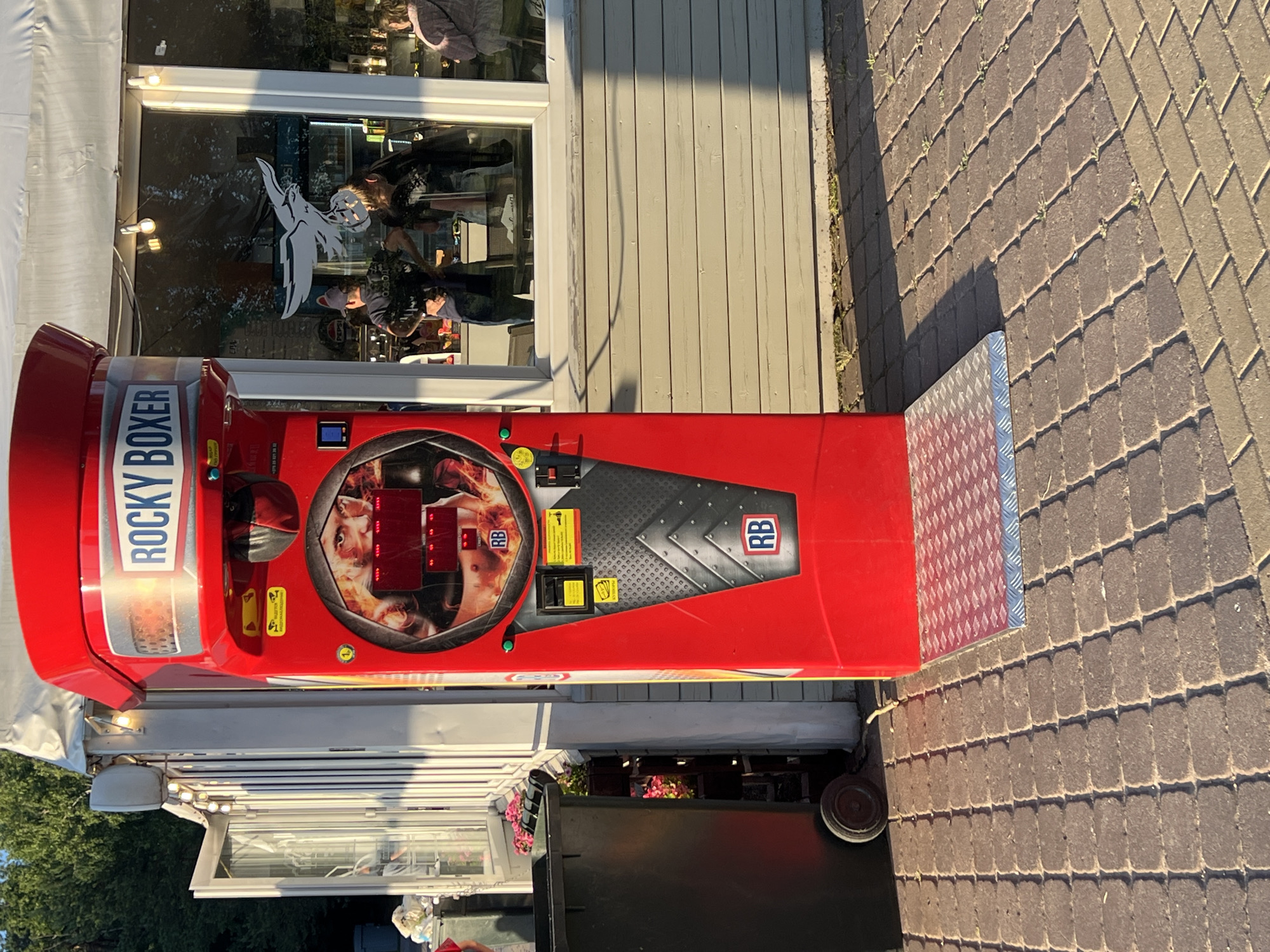 The image shows a red arcade boxing machine, labeled "ROCKY BOXER," situated outdoors on a paved walkway. The machine features a screen displaying a boxer's face and digital readouts. A metal platform is in front of the machine. To the left of the boxing machine is a large, dark-colored trash bin with wheels. In the background, there is a building with large windows, through which people can be seen inside. The scene appears to be in a public area, possibly a park or a street in Minsk, Belarus, given the typical appearance of such arcade machines and the environment. The lighting suggests it is daytime, with shadows cast on the ground.