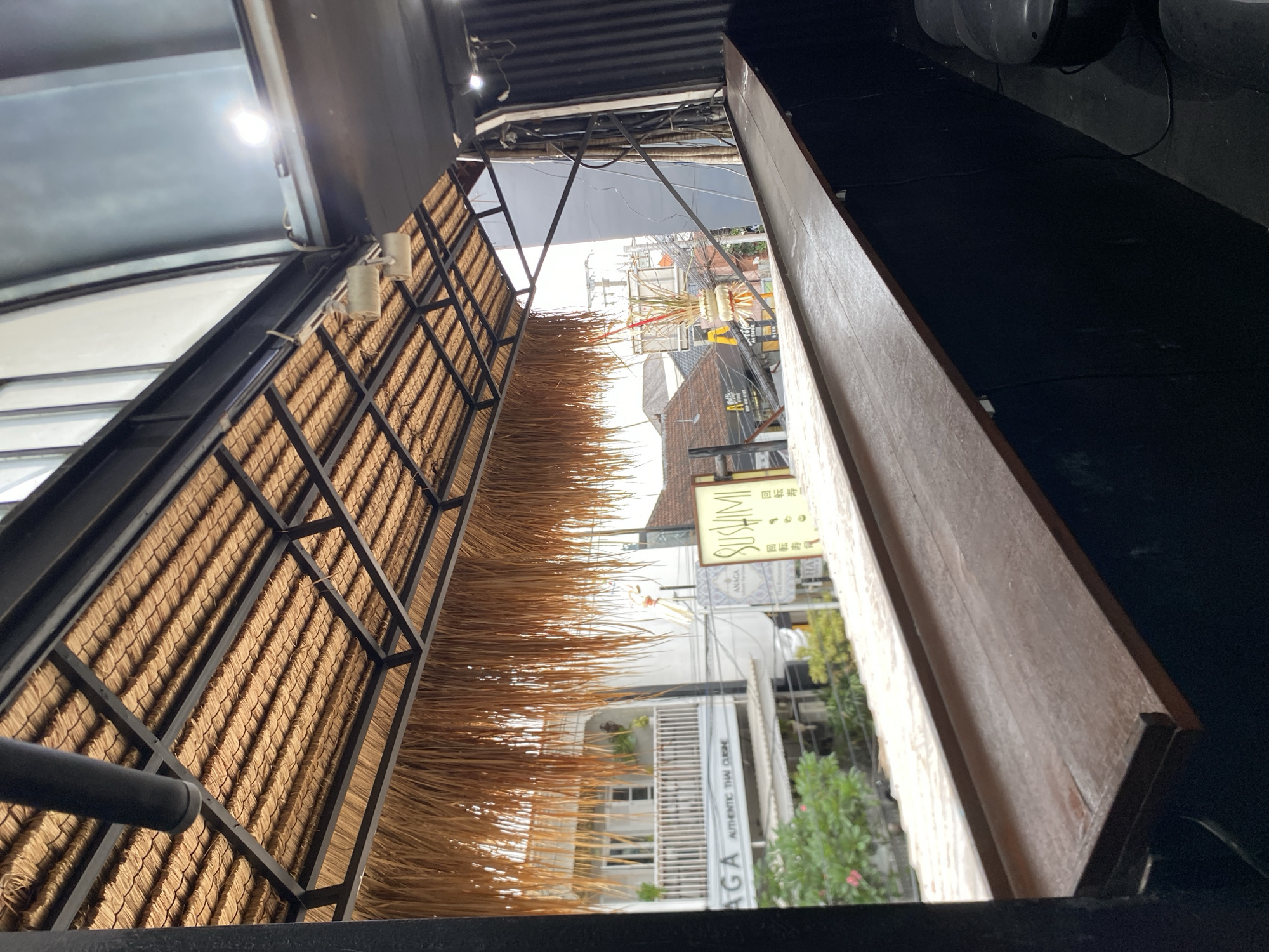 This image shows a view from a thatched-roof covered outdoor seating area, likely a restaurant or bar, looking out onto a street in North Kuta, Indonesia. In the foreground, a dark wooden counter or bar top is visible, extending across the frame. Above the counter, a thatched roof supported by a black metal frame is seen. Hanging from the roof are dried palm fronds, creating a naturalistic aesthetic.
Beyond the seating area, a street scene unfolds. A building with white balconies and windows is prominent on the left, with text visible on its facade that reads "ANAGA AUTHENTIC THAI CUISINE." To the right of this building, a sign with the word "SUSHI" in yellow and black lettering is displayed, along with some Japanese characters. Further down the street, other buildings with tiled roofs and various signs are visible, suggesting a commercial area. There are also power lines and utility poles. The overall impression is of a tropical, bustling urban environment. No people are clearly visible in the image.