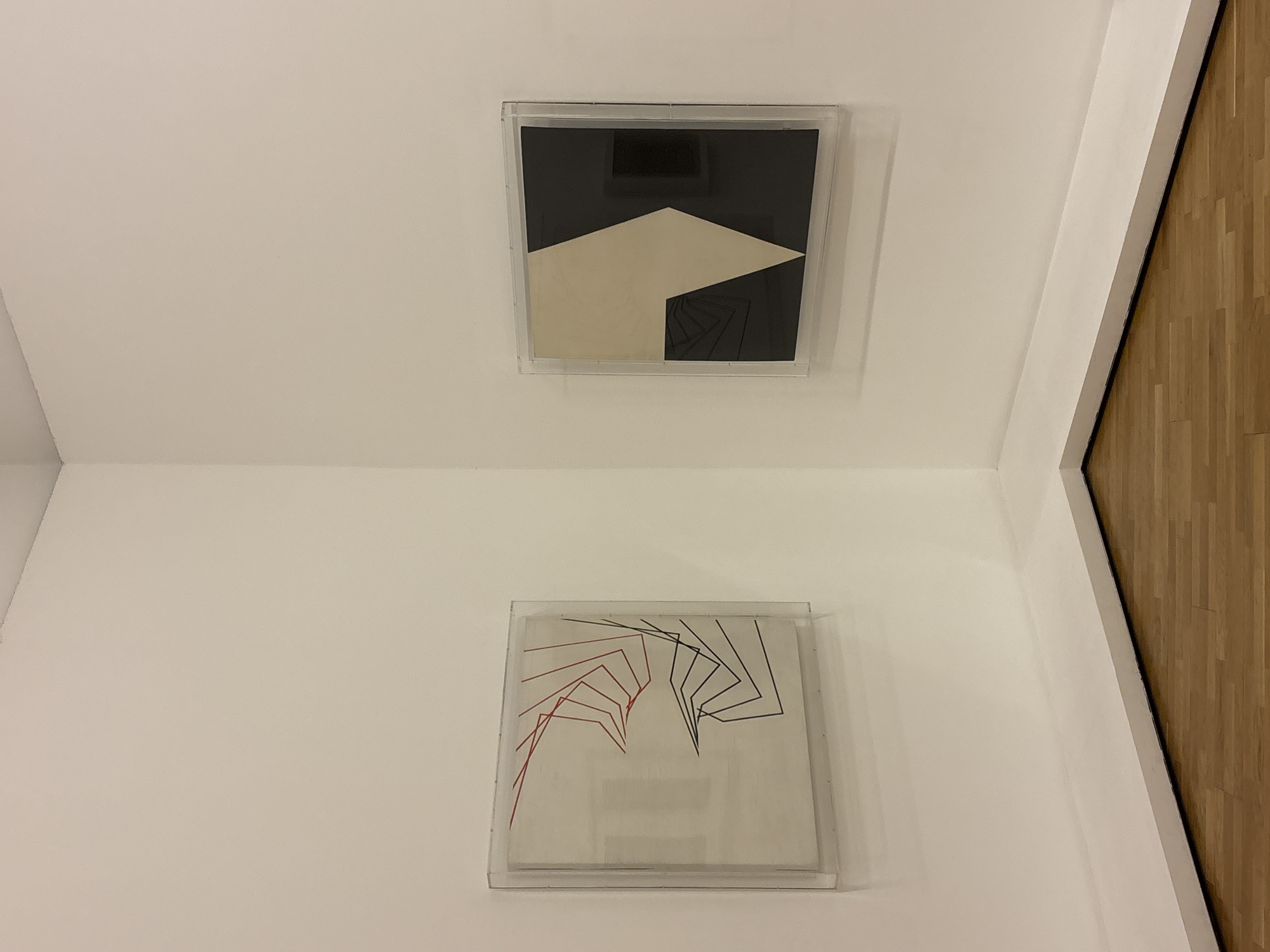 The image depicts two pieces of framed artwork mounted on a white wall within what appears to be an interior space. The artwork on the top is a black and white abstract composition, while the lower artwork features red and black lines converging in the center. To the right of the artworks, wood flooring is visible, suggesting the edge of a room or corridor. The image was taken in São Paulo, Brazil.