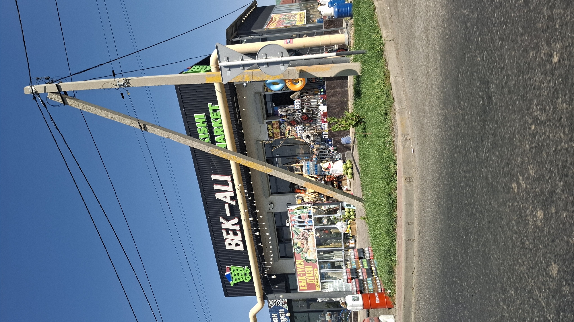 The image shows a street-level view of a commercial establishment in Shymkent, Kazakhstan. The building, identified by prominent signage as "BEK-ALI MARKET," features a black facade with illuminated lettering. In front of the building, various items are displayed, including stacks of bottled drinks, watermelons, tools, and other merchandise. A concrete utility pole, angled and supporting electrical wires, stands in the foreground, partially obscuring the storefront. The ground is paved with asphalt, and a strip of green grass borders the sidewalk in front of the market. The sky is clear and blue, indicating a sunny day. There are no visible people actively engaged in transactions or identifiable actions within the frame.