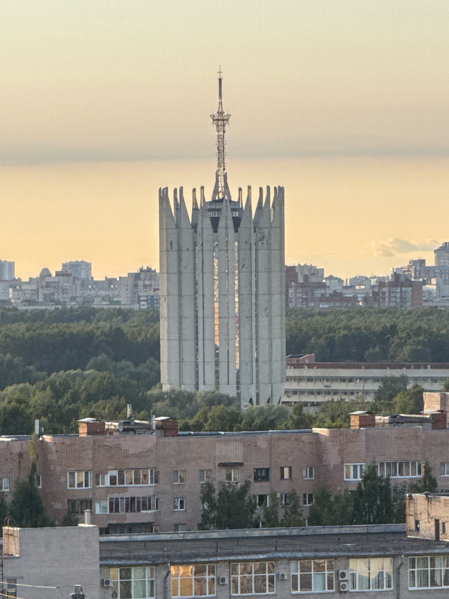 This is a high-angle, long shot photograph of a city skyline in Unknown, Russia, during twilight. The foreground features a multi-story apartment building constructed of brick, with numerous windows and air conditioning units visible. Behind it, another, lower apartment building is partially visible. Beyond these residential structures, a dense forest of green trees dominates the mid-ground.
In the background, a distinctive, tall, white, Brutalist-style tower stands prominently, resembling a stylized crown or a pointed flame. This tower is topped with a complex antenna structure. To the left of this tower, several other city buildings and apartment blocks are visible, suggesting a densely populated urban area. The sky is a soft gradient of pale orange and yellow, indicative of sunset or sunrise. The overall atmosphere is serene and urban.