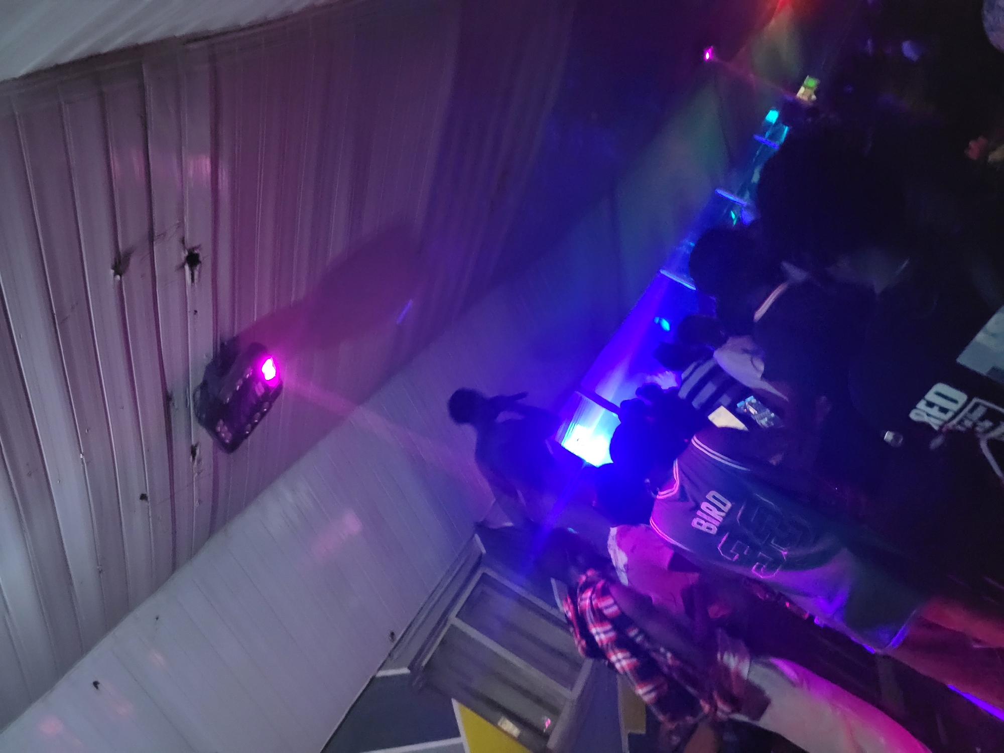 The image depicts an indoor gathering with several individuals present, illuminated by purple and blue stage lighting. A projector is mounted on the ceiling, casting beams of light. In the foreground, a person is wearing a basketball jersey with the name "BIRD" and the number "33" on the back. Other individuals are visible in the background, some appearing to be dancing or socializing. The environment suggests a nightlife setting, possibly a club or event space. The location is identified as Agwala, Nigeria.