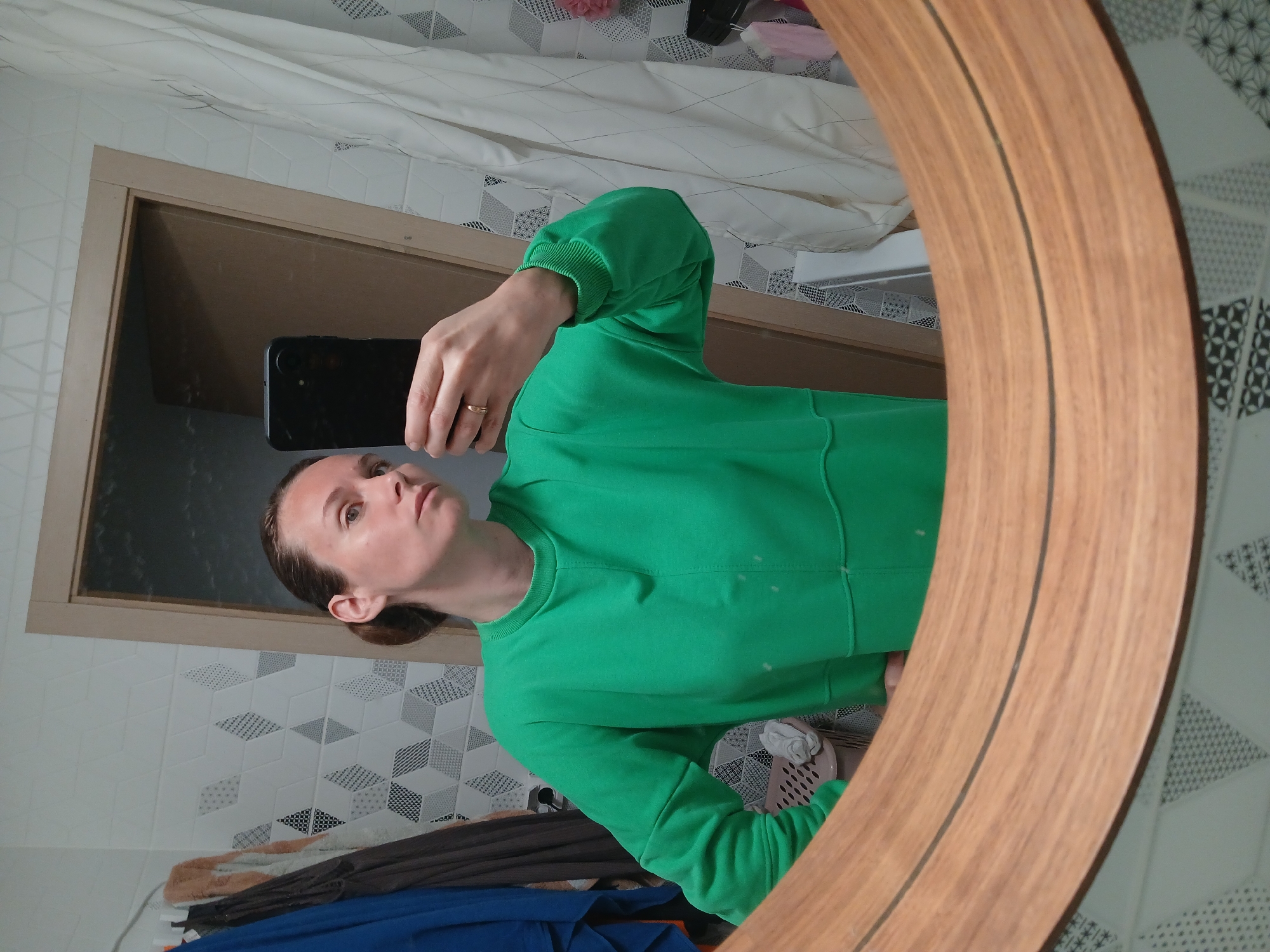 A woman is taking a mirror selfie in a bathroom. She is wearing a bright green sweatshirt and has her hair pulled back. She is holding a black smartphone in her right hand. The bathroom has white patterned tiles on the wall and a wooden-framed mirror. There are clothes hanging on a rack to the left of the woman and a white shower curtain on the right. The scene is indoors and appears to be in a residential setting.