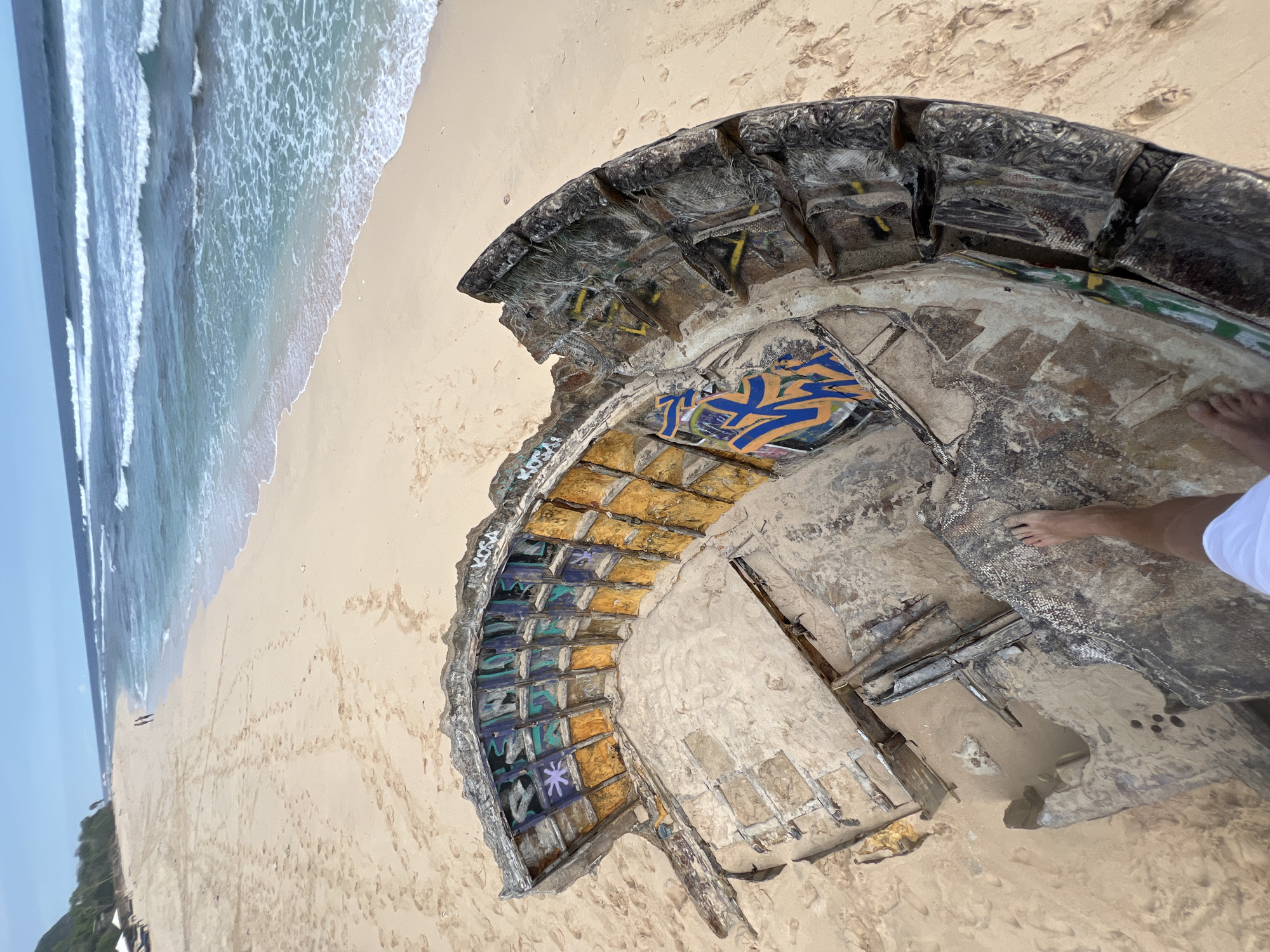A half-circle structure stands on a sandy beach. The structure's interior is composed of tiled sections, some painted with graffiti. The exterior is weathered and grey, exhibiting signs of decay. The ocean is visible in the background, with waves breaking on the shore. Two people are visible in the background, walking on the beach. A person's bare feet and a portion of white clothing are visible within the structure. The location is Pecatu, Indonesia.