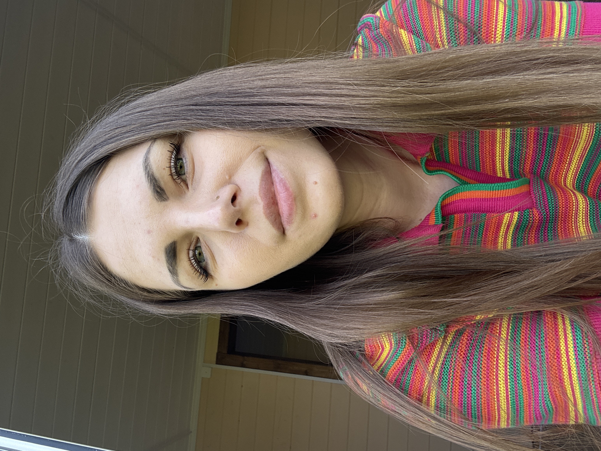 A woman is pictured indoors in a vertical, medium shot. She has long brown hair, fair skin, and green eyes. She is wearing a brightly colored striped top with pink, green, yellow, and orange. The background shows a beige wall and a window with a dark brown frame. The location is assumed to be Razdolye, Russia, but there are no visual cues in the image that confirm this location.
