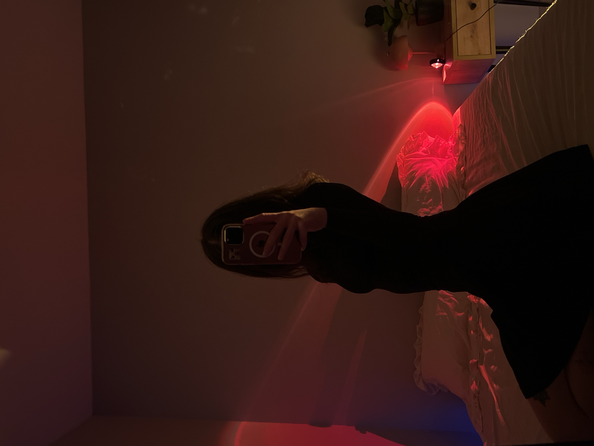 A person is taking a selfie in a dimly lit bedroom. The person is wearing a black dress and is holding a smartphone in their right hand. The smartphone is displaying a pinkish-red light effect. The background of the image shows a bed with white sheets, a nightstand with a lamp and a plant, and a wall with red and blue light projections. The location is not Ternopil, Ukraine.