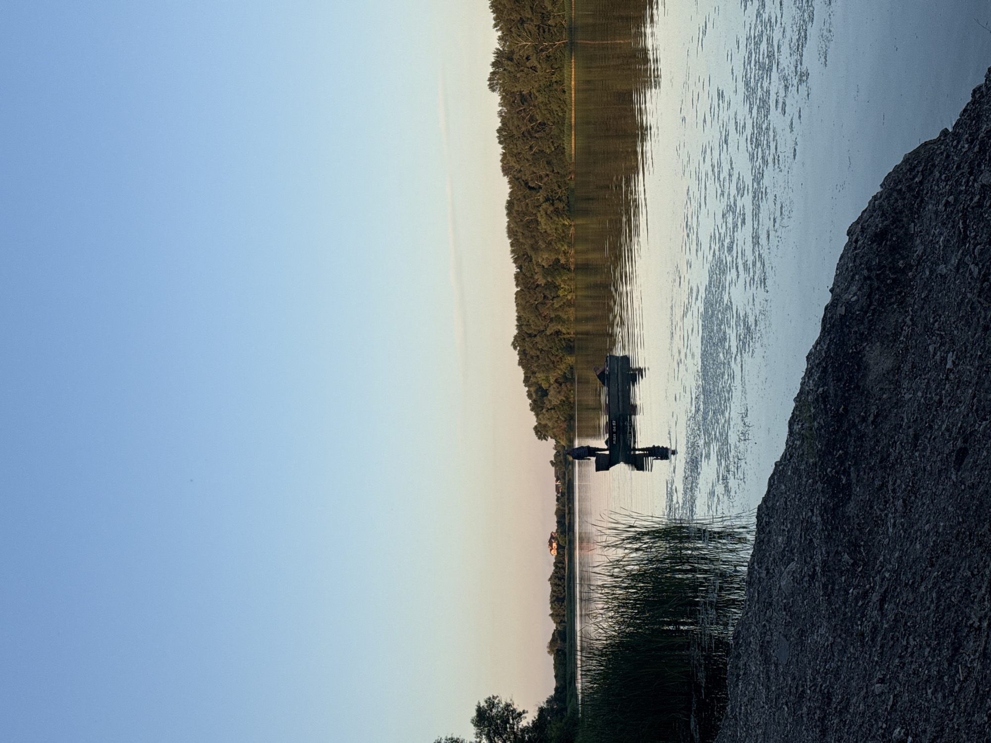 In the city of Dnipro, Ukraine, this image captures a serene sunset scene on the Dnipro River. A lone fisherman stands in a small boat anchored in the calm water. The sky transitions from a soft blue to a pale orange near the horizon, reflecting on the water's surface. Dense green trees line the opposite bank, with a few houses visible in the distance. Reeds grow tall on the left bank, partially obscuring the water's edge, which is covered in dark gravel. The overall atmosphere is peaceful, with the natural beauty of the river setting a tranquil mood.