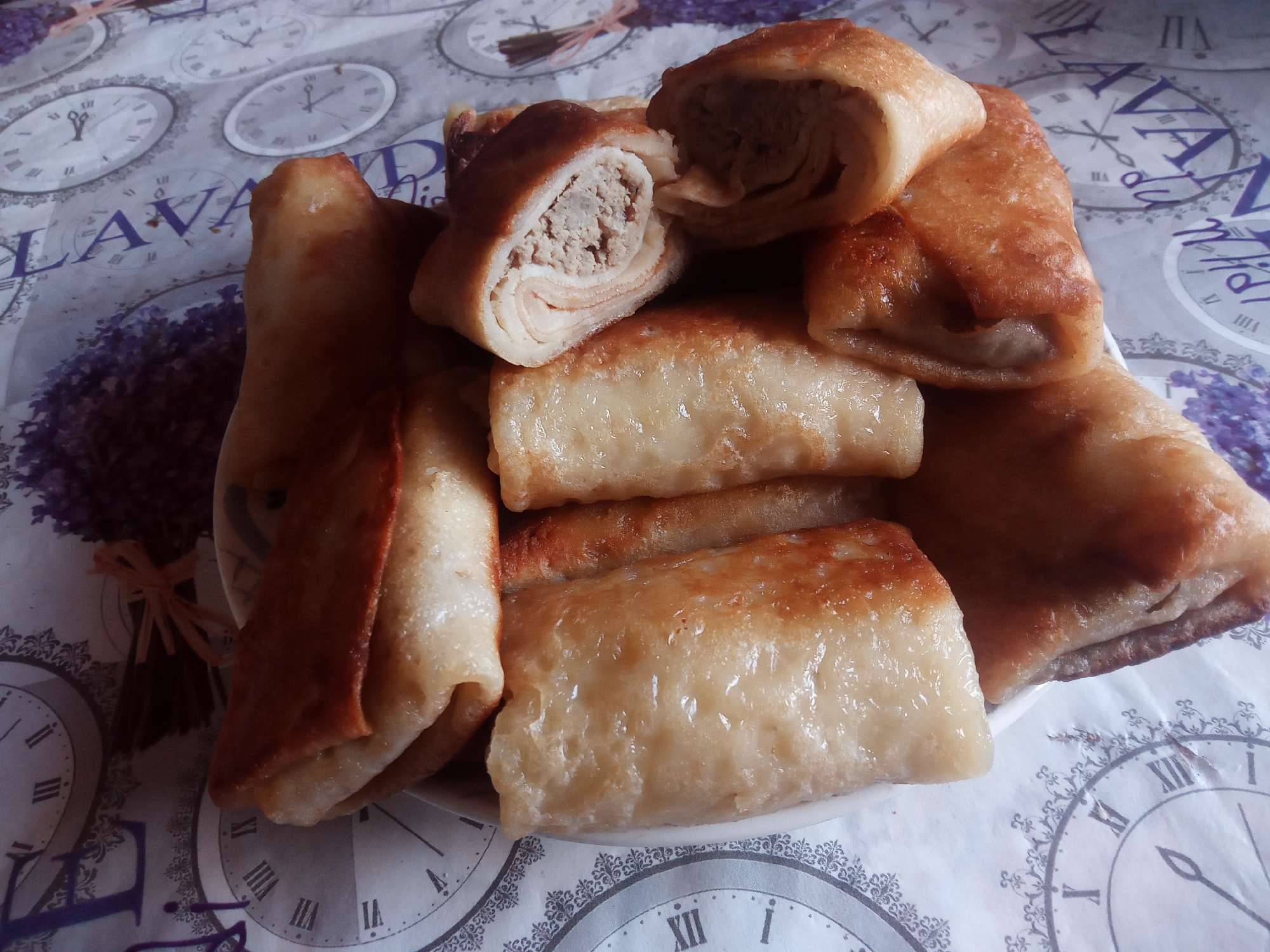 A close-up, high-angle shot shows a white plate filled with golden-brown crepes, or blini, filled with a light-colored meat mixture. The crepes are arranged in a pile, with some cut open to reveal the filling. The plate sits on a tablecloth with a lavender and clock pattern. The image does not contain any people, nor does it provide information about the city of Usman, Russia.