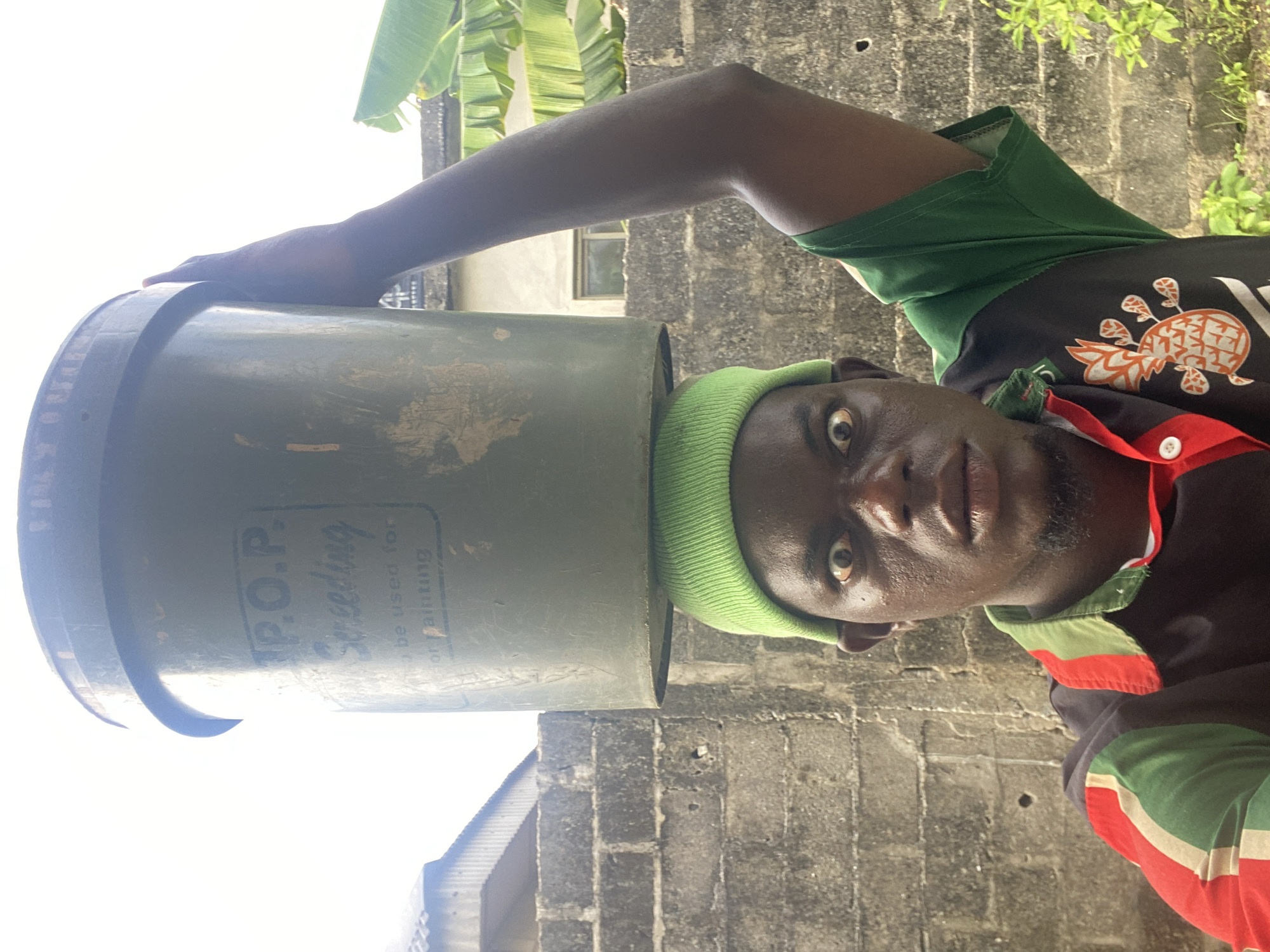 A man wearing a green beanie and a black, green, and red shirt with a pineapple design on the front is holding a large, green bucket labeled "P.O.P. Speeding" on his head. He is looking directly at the camera with wide eyes. The background includes a gray brick wall, greenery, and a building with a white roof and a window. The location is Unknown, Unknown.