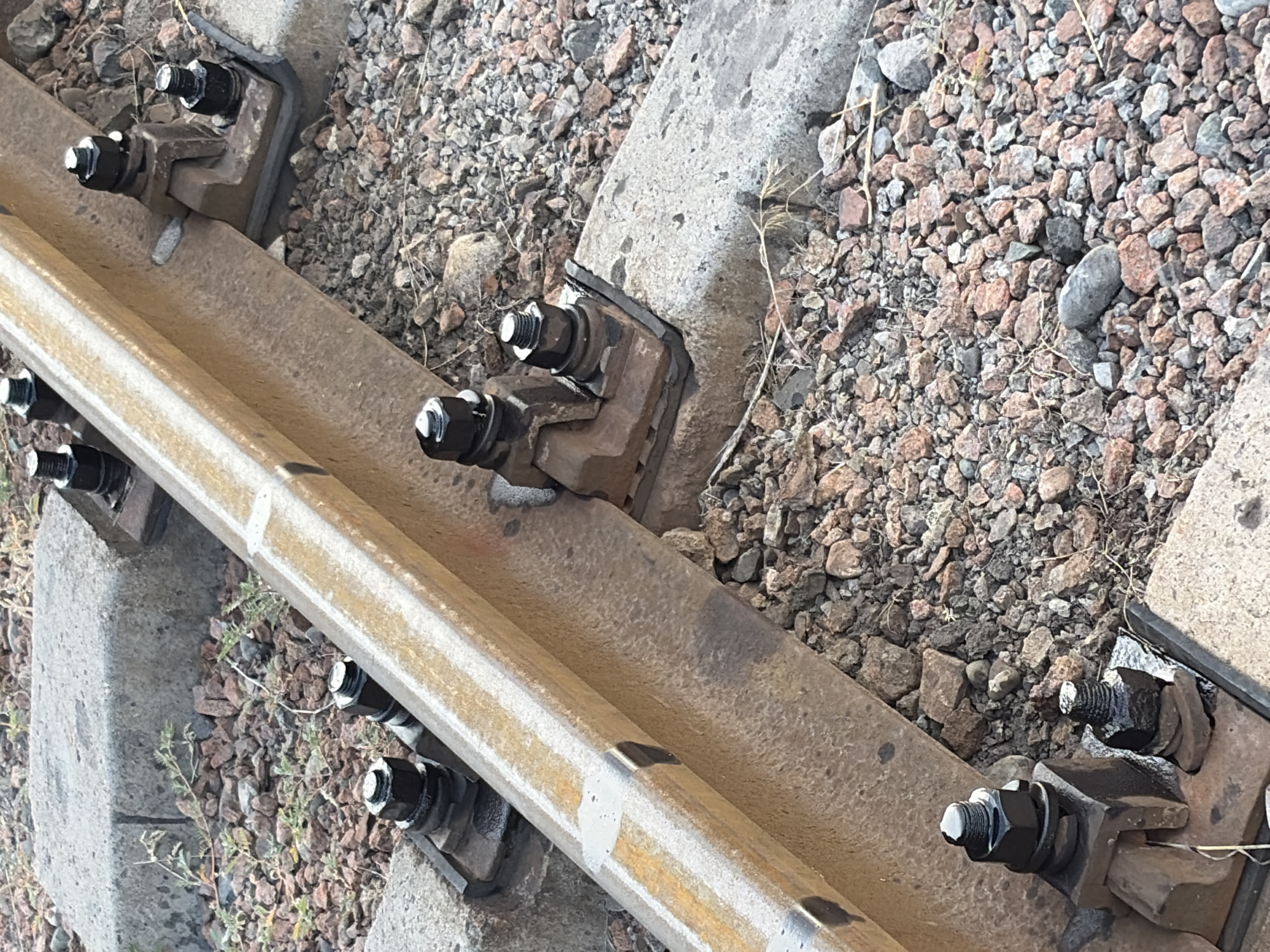 The image shows a close-up view of a railway track, focusing on the fasteners that secure the rail to concrete sleepers. The subjects are the railway components themselves: the steel rail, concrete sleepers, and the metal clips and bolts that hold them together. The scene is a section of a railway line, with ballast stones surrounding the sleepers.
There is no discernible activity or event taking place; the image appears to be a static capture of the infrastructure.
Notable details include the texture of the weathered steel rail, the rough surface of the concrete sleepers, and the black painted bolts and nuts. The ballast is composed of small, multi-colored stones, predominantly reddish-brown and gray. The lighting suggests it is daytime, with no strong shadows indicating a specific time of day. There are no clear location cues beyond it being a railway, other than the provided context of Kant, Kyrgyzstan. No emotions are conveyed.
There is no visible text in the image.