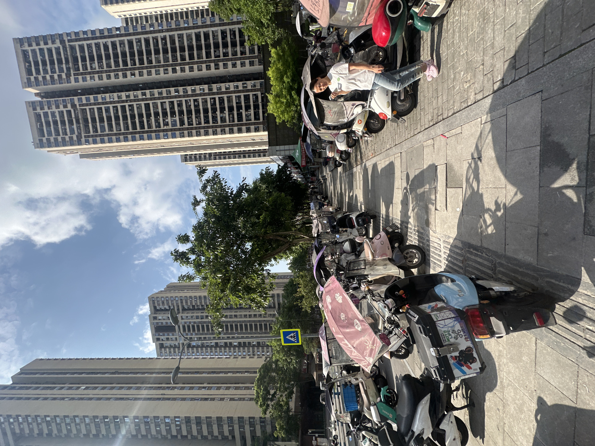 In an urban environment consistent with the Chongqing High-tech Zone, China, a row of parked electric scooters and tricycles lines a paved sidewalk. The scooters, many with colorful canopies, are arranged in a dense formation. A woman in a white t-shirt and blue jeans, wearing pink Crocs, walks along the sidewalk. She is looking at her phone, which she holds in her left hand. The background is dominated by tall, modern apartment buildings, characteristic of a dense city. Sunlight casts shadows across the pavement, indicating a clear day. A pedestrian crossing sign is visible further down the sidewalk, and trees are interspersed among the buildings and scooters. The scene depicts a typical urban streetscape with a focus on personal transportation options.