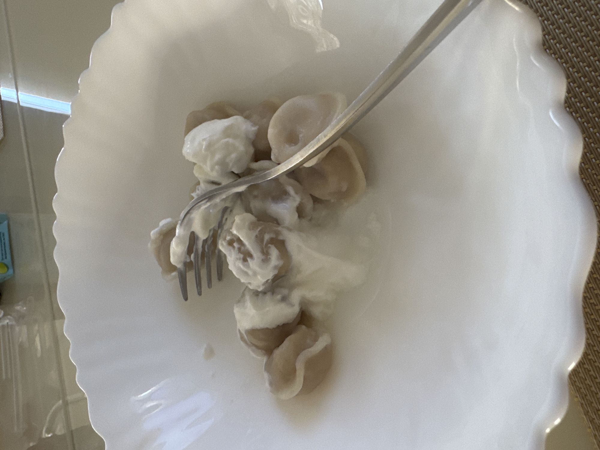 A close-up, eye-level shot shows a white, scalloped bowl filled with pelmeni, a type of dumpling, covered in a generous dollop of sour cream. A silver fork is partially submerged in the food, with its tines picking up some of the dumplings and cream. The bowl sits on a woven placemat. The background is blurred, but appears to be a table surface with reflections. The lighting suggests an indoor setting, possibly a kitchen or dining room, with the natural light filtering in from a window. The image does not provide any specific details or context to locate it in Uyutnoye, Ukraine.