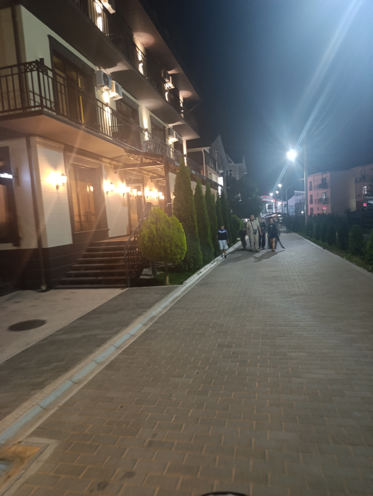 The image depicts a nighttime scene on a paved walkway in Bosteri, Kyrgyzstan. To the left, a multi-story building with balconies and illuminated windows is visible. Lights are also visible on the exterior of the building, and air conditioning units are mounted on the facade. A set of stairs leads up to the building's entrance, flanked by a decorative bush.
A group of people, appearing to be about five in number, are walking along the pathway away from the camera. They are silhouetted against the dim lighting. Further down the walkway, more trees are planted in rows along the side. In the background, other buildings are visible, some with illuminated windows, and a streetlamp shines brightly in the distance, casting lens flares. The walkway itself is constructed of brick pavers. The overall atmosphere is that of a public space at night, possibly a resort area given the presence of the building and manicured landscaping.