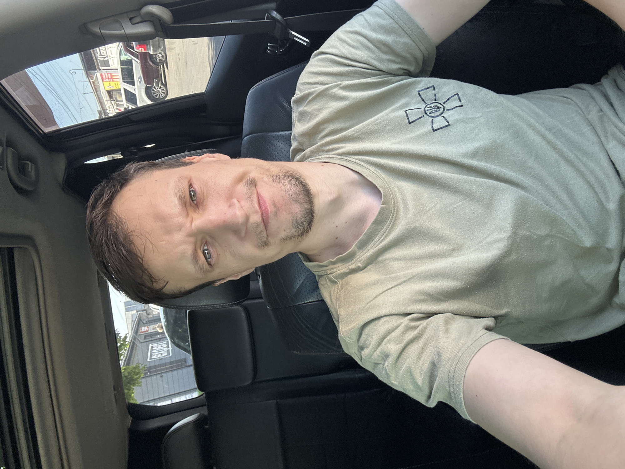 A man with short brown hair and a light beard is in the driver's seat of a car, taking a selfie. He is wearing a light green t-shirt with a dark emblem on the left chest. The t-shirt appears to be made of a lightweight cotton material, and the emblem consists of a stylized cross with a shield in the center. The man's eyes are looking directly at the camera, and he has a slight smile on his face. The interior of the car is visible behind him, including black leather seats and a sunroof. The background outside the car windows shows a street scene with buildings and parked cars, suggesting an urban environment. The scene appears to be set in a city, possibly in Ukraine given the emblem on the man's shirt which resembles a Ukrainian military insignia. Without additional information or context, it is not possible to definitively identify the specific location in Kamianets-Podilskyi, Ukraine.
