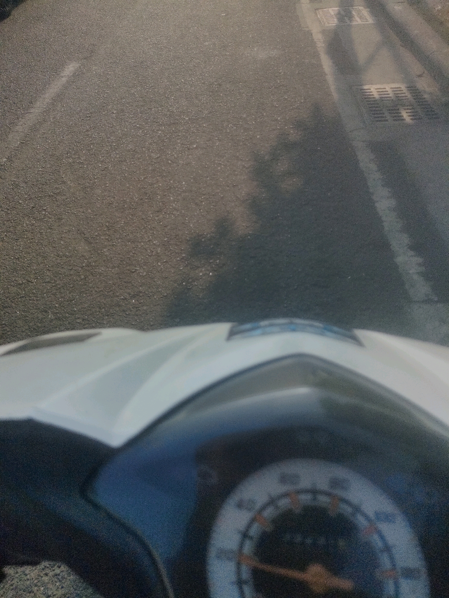 This is a first-person perspective, forward-facing, slightly high-angle, medium shot captured from a motorcycle in Surabaya, Indonesia. The image focuses on the road ahead and the motorcycle's front panel and dashboard, which is blurred. The road is asphalt with visible texture and some lighter markings, possibly from previous paint. To the right of the road, there's a concrete drainage grate with a shadow cast over it. The shadow appears to be from overhead lines or structures, and possibly a person or object. The lighting suggests it is daytime, with sunlight casting shadows. The overall scene depicts a typical urban road environment in Surabaya.